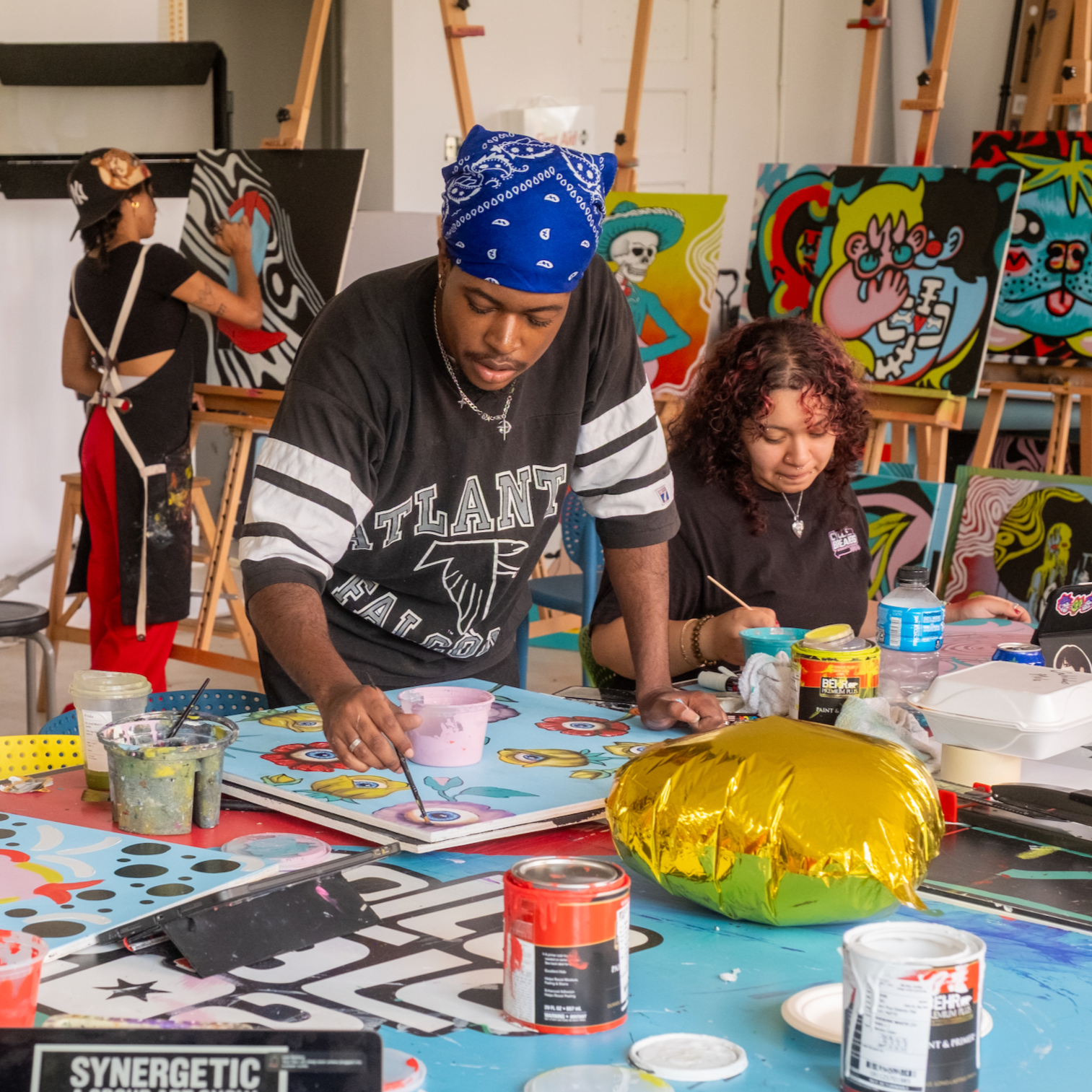 Three young people work on various art pieces in a studio. One person works on a canvas piece on an easel. The two people in the foreground work on other pieces at a table.