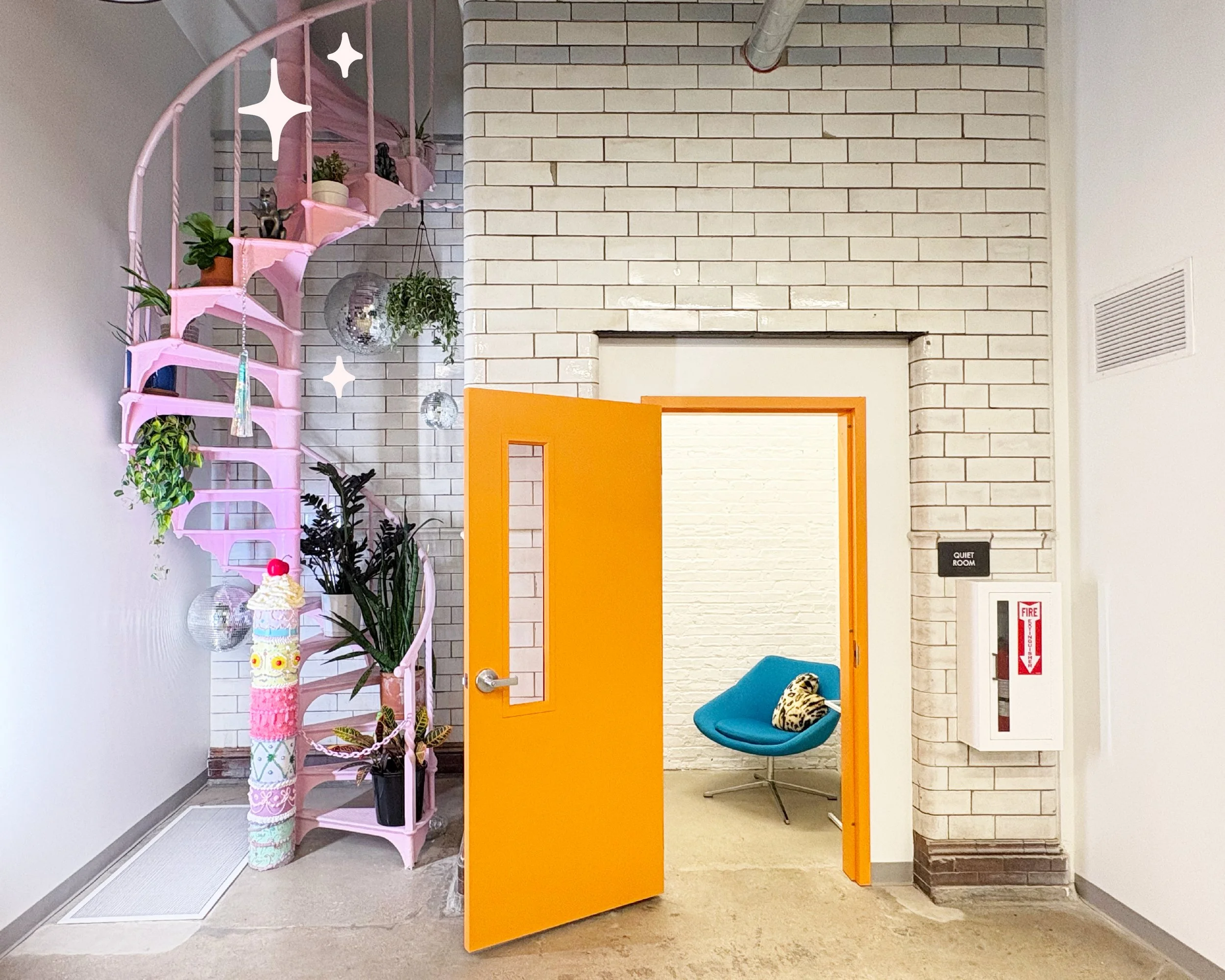 A closer look at the "Main Event Space" pink spiral staircase, which is lined with plants along the steps. A "Quiet Room" is visible to the righ tof the stairs. Inside the room, sits a blue lounge  chair. A fire extinguisher is on the wall nearby.