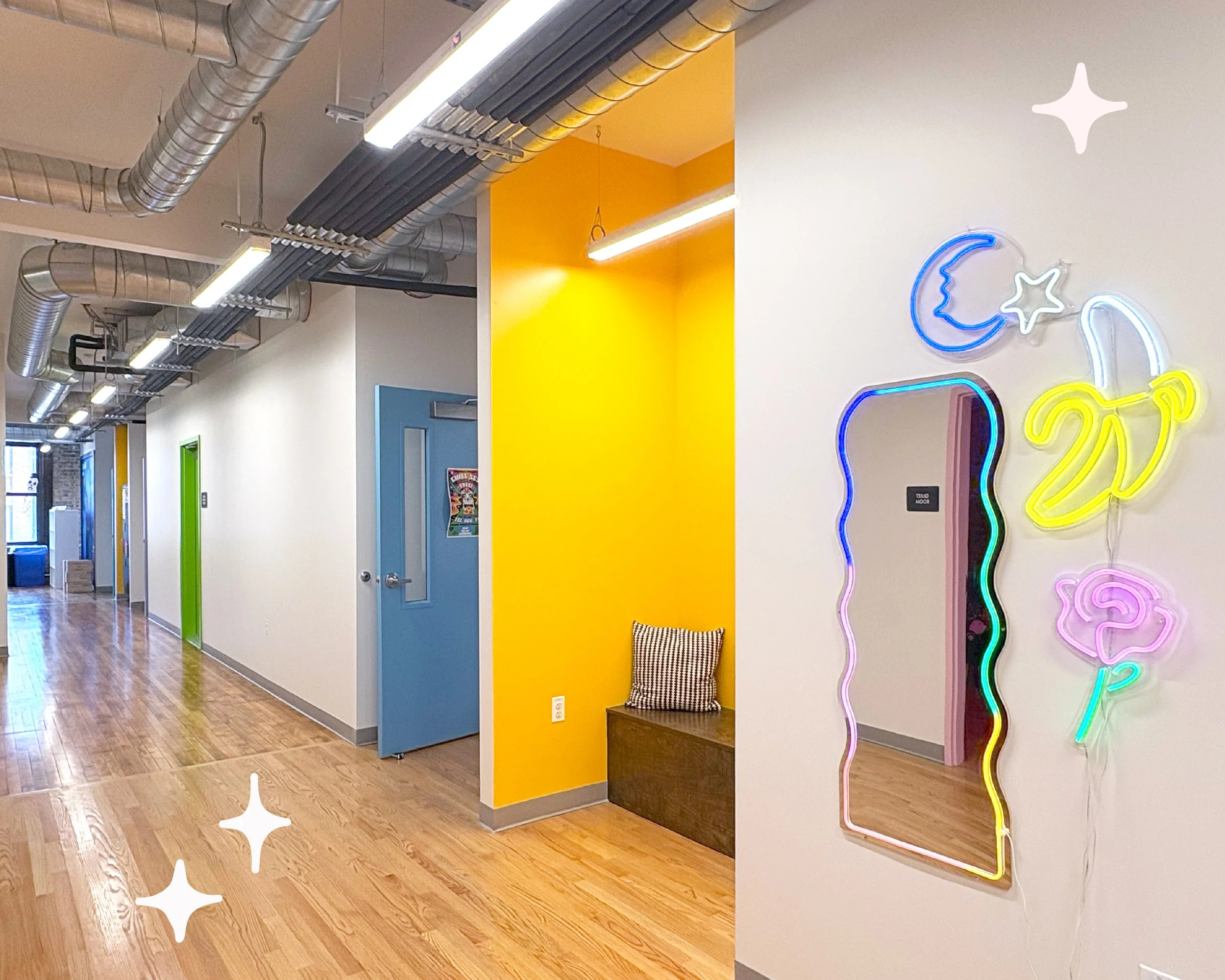 View of a halway with wood floors. A yellow nook is visible, as well as several colorful doors down the hallway. In the foreground, neon wall decor, and a mirror are hung.