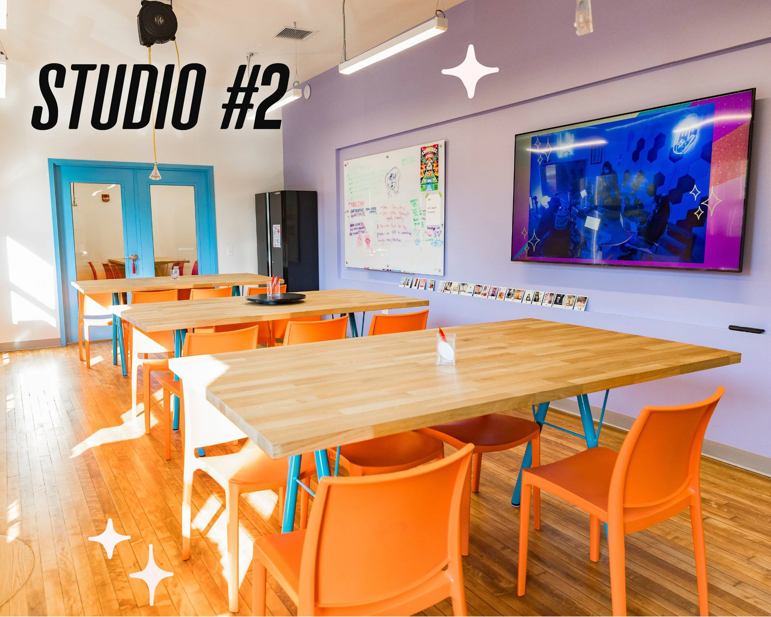 "Studio #2" reads the graphic text over another bright workspace. Multiple tables with orange chairs are in the foreground. A lavender accent wall with a video screen and a white board are in the background.