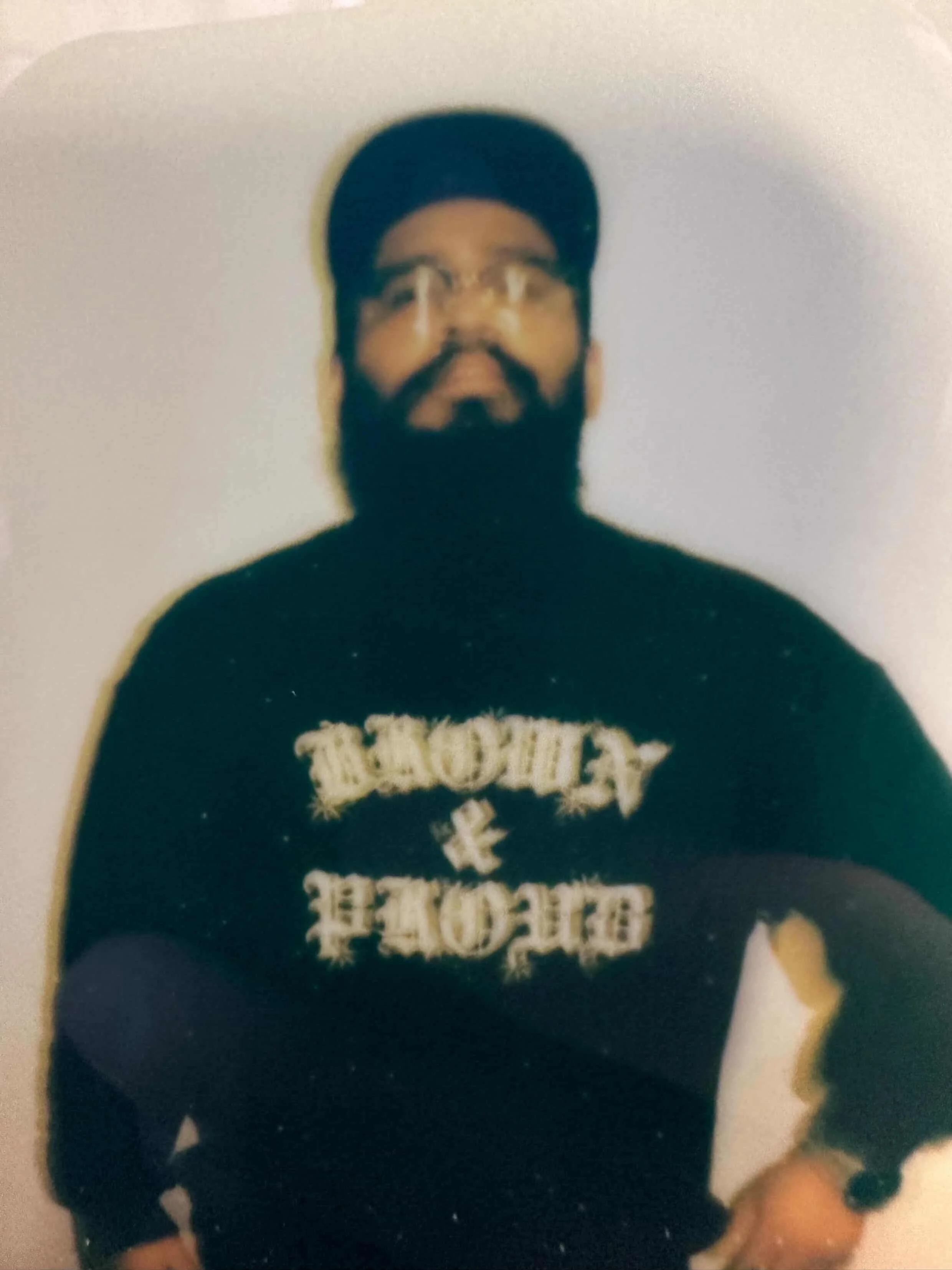 A person with a beard, glasses, and a baseball hat poses in front of a white wall. This photo of them is sylized and slighly blurry. They are wearing a sweatshirt that reads, "brown & proud."