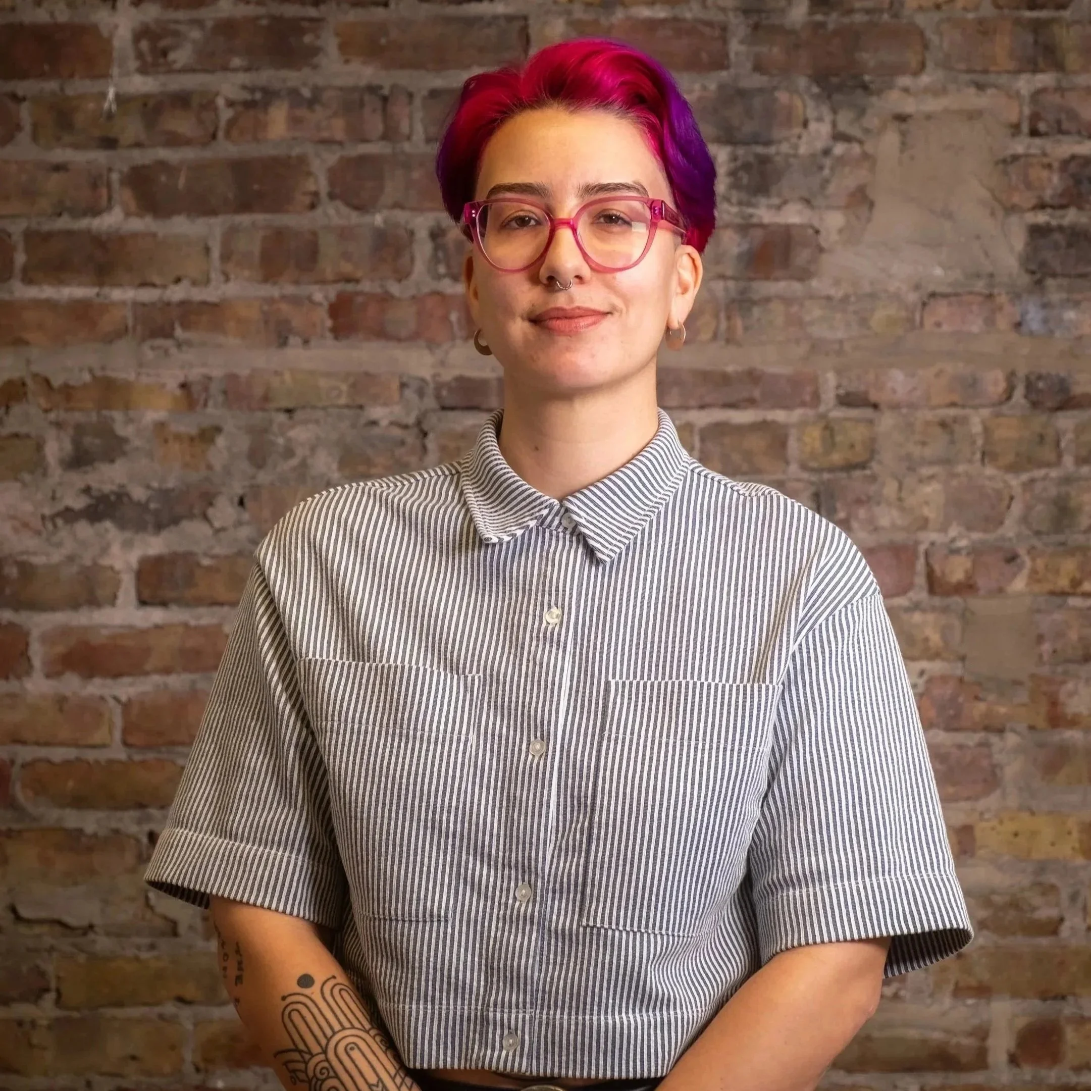 A person with short fuchsia hair poses in front of a brick wall. They are wearing a pinstripe button-up shirt and a colorful pair of glasses.