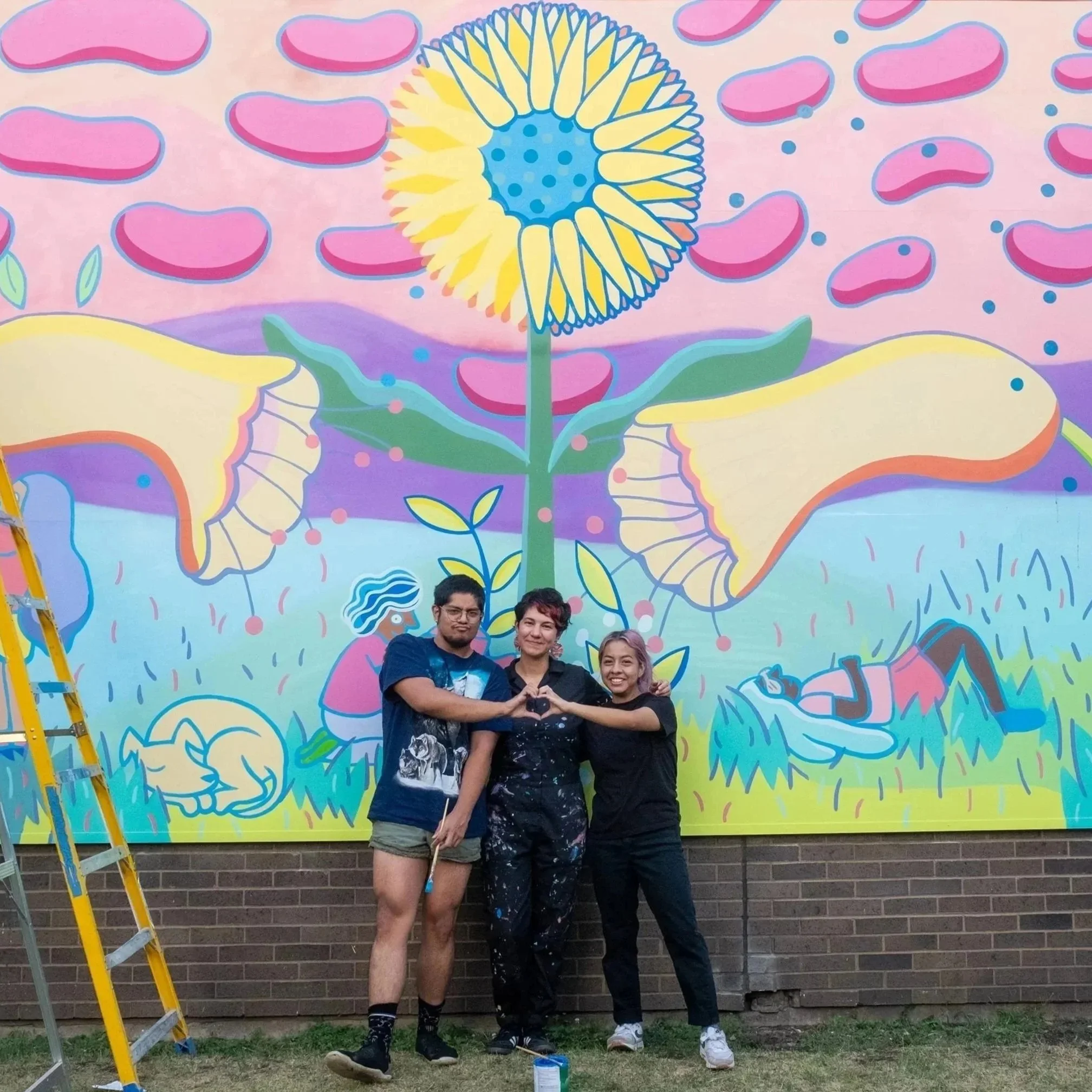 Three young people pose in front of amural that features a dandelion, and other stylized flowers. The person in the center wears painters overalls, and the two people on either side lean over to each other to form a heart with their hands.