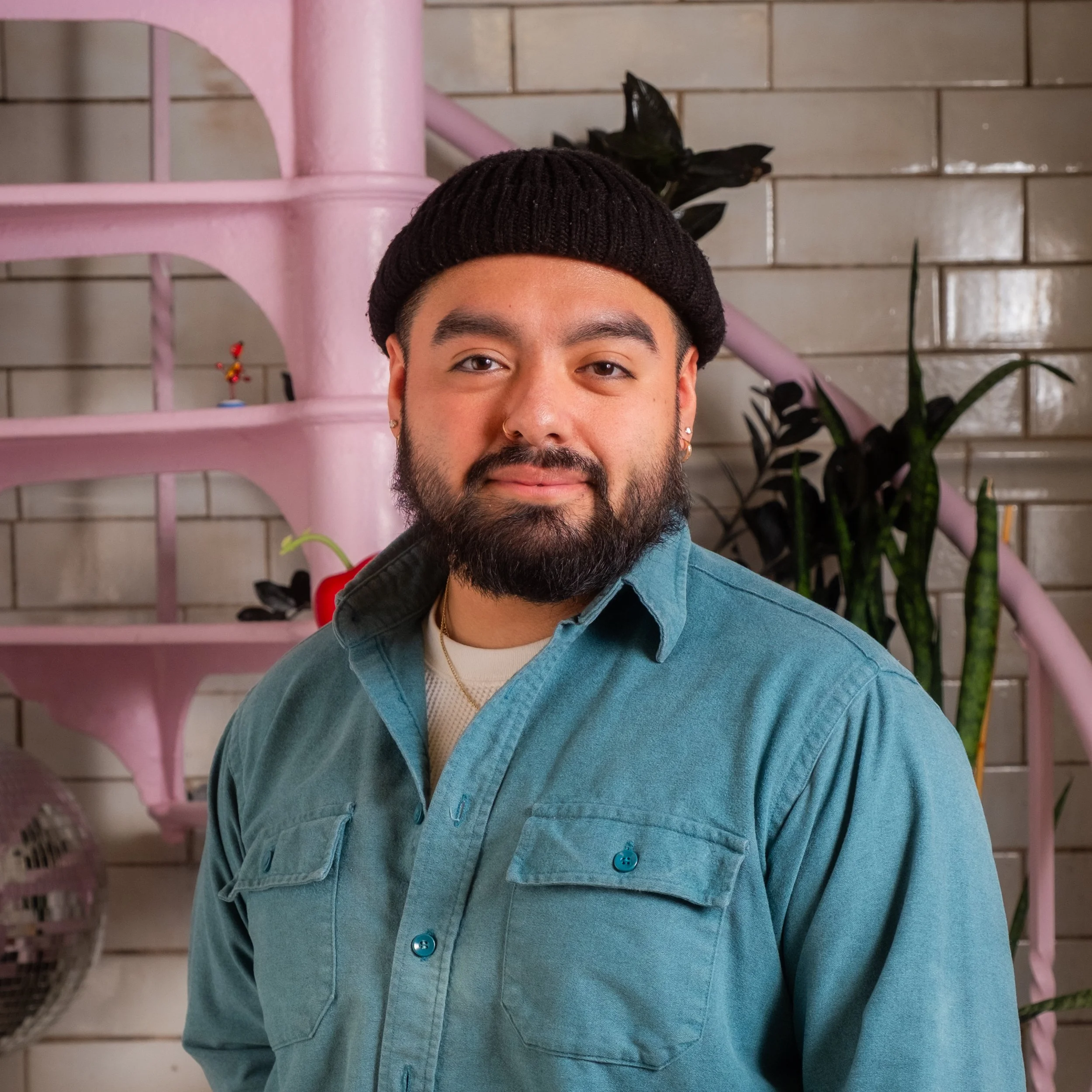 A person with a trimmed beard poses in front of a pink spiral staircase. They are wearing a rolled-up black beanie and a green button-up shirt.