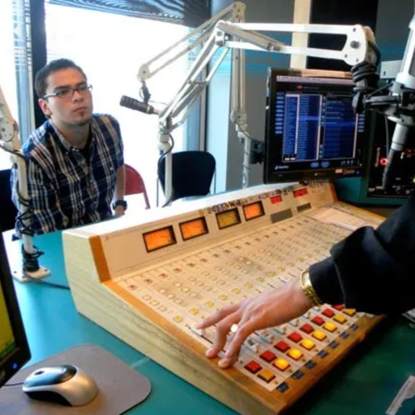 A young person with short hair, glasses, and a flannel shirt sits in a radio studio. In the foreground hands hover over an audio mixing board.