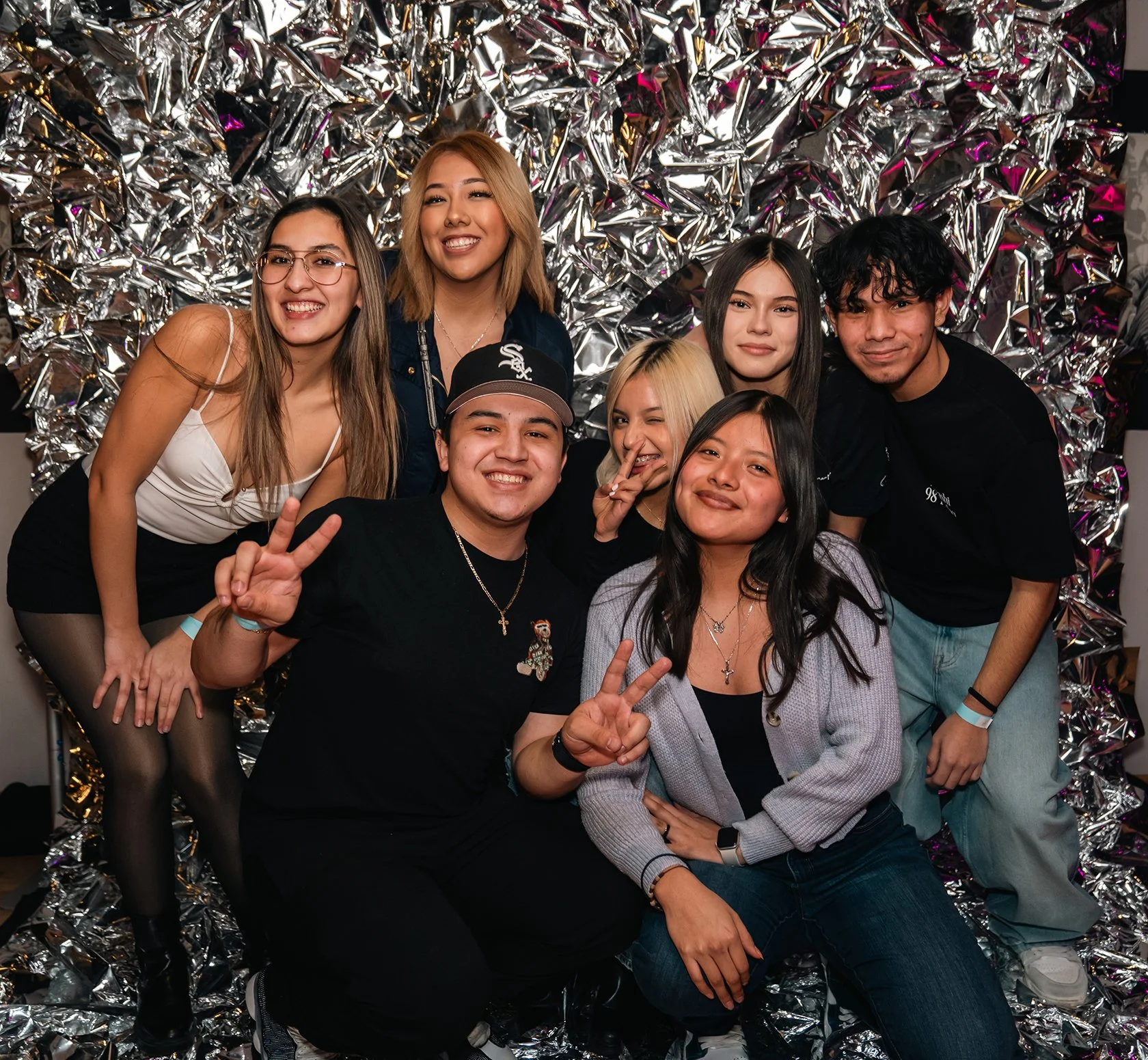A group of seven young people pose in front of a silver metalic background. They are smiling and posing in a group.