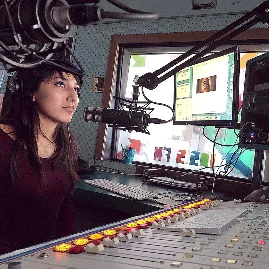 A young person sits in a radio booth. Various monitors, soundboards, and microphones are visible.
