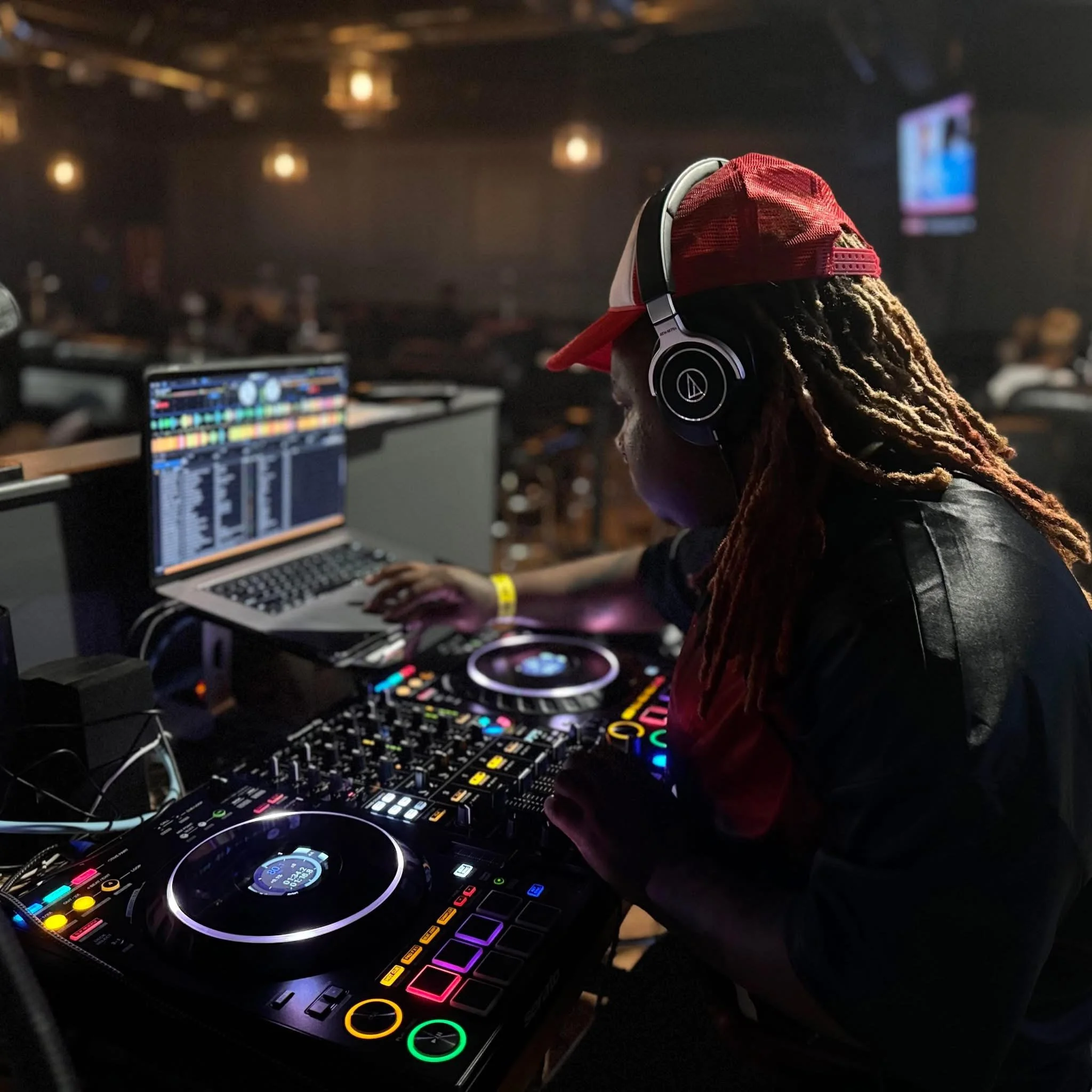 A person with locks and a baseball hat faces partially away. They are wearing wired headphone and use a comuter to navigate music software. The table in front of them is covered in turntables andsound mixers. They are performing a DJ set.
