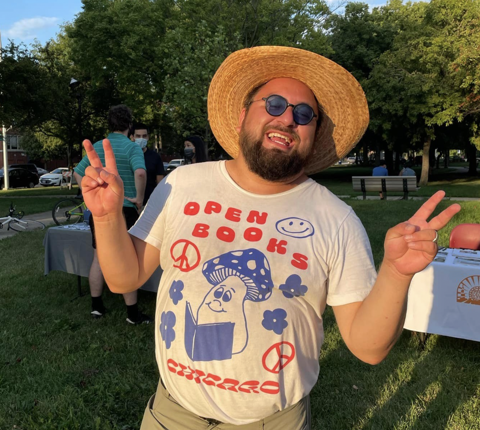 A person with a beard poses wearing a straw hat and tinted glasses. They are smiling and are using their hands to throw up peace signs. They are in a park at sunset.