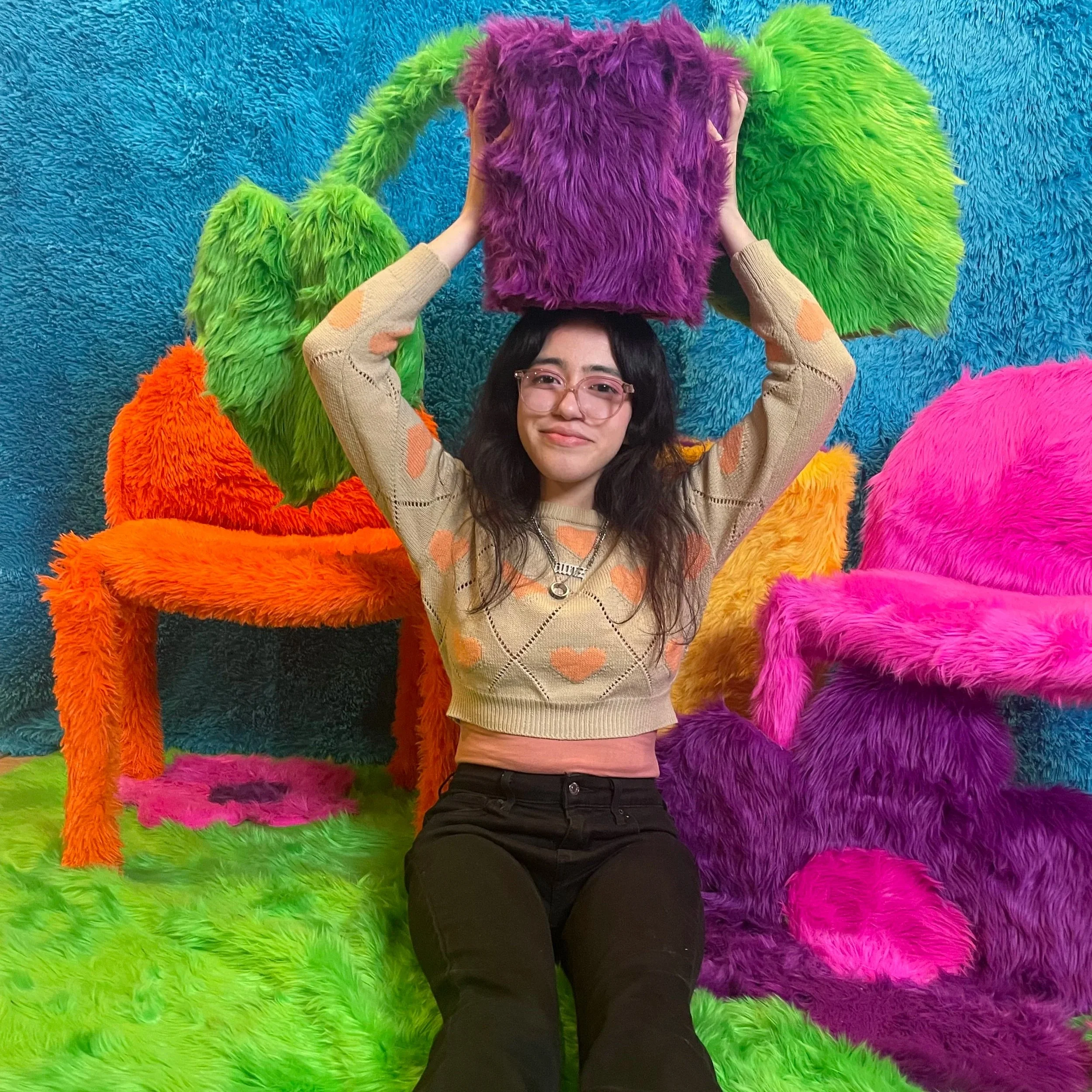 A young person posing in front of a fuzzy multi-colored background. They are raising a fuzzy purple block over their head.