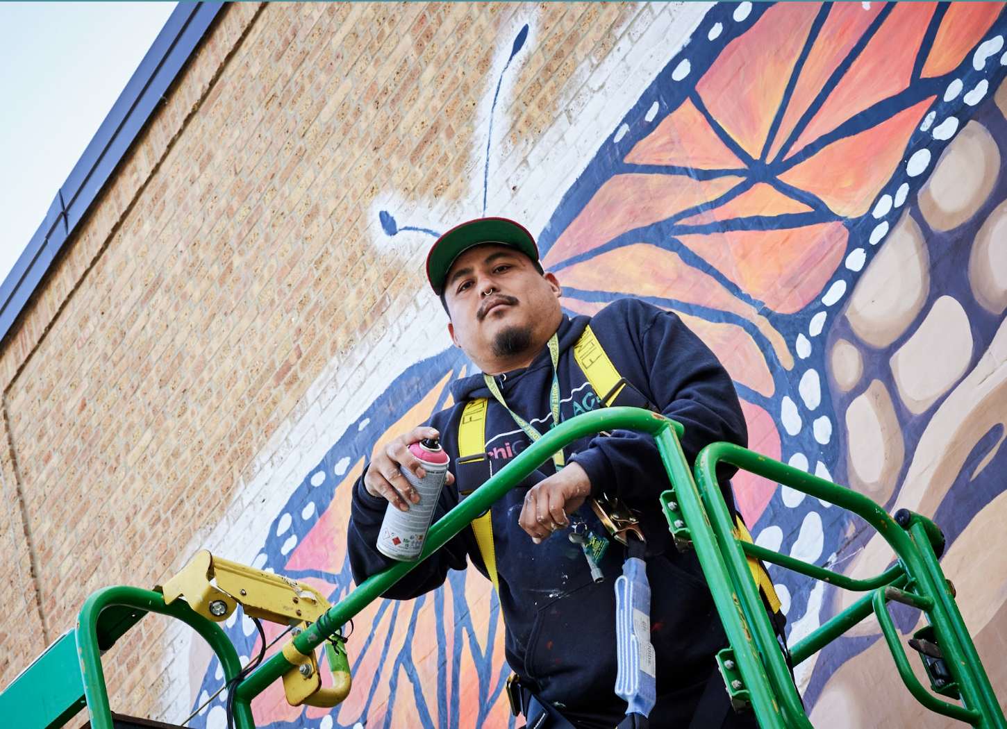 A person wearing a black hoodie and a baseball hat looks down from a lift. They are holding a can of spray paint, and behind them is a monarch butterfly mural painted on a brick wall. The angle makes it seem like the person on the lift has antennas.