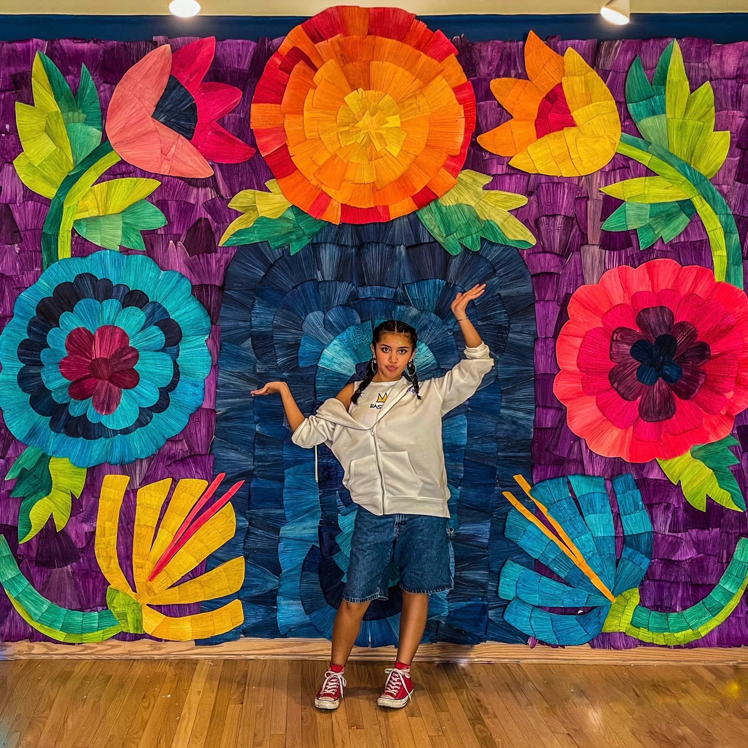 A young person poses with their arms outstreatched in front of a colorful art installation featuring multicolored flowers and a deep purple background.