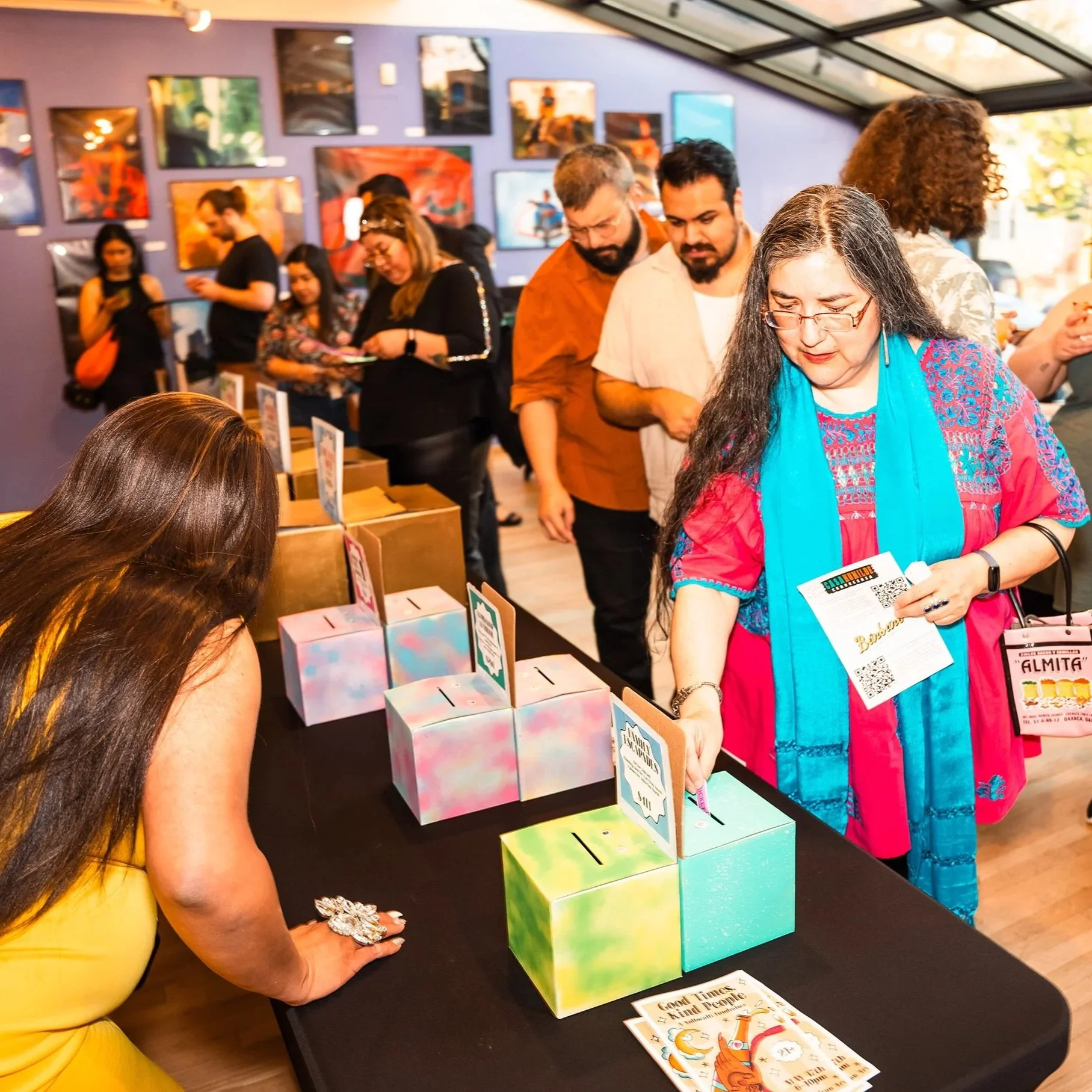 A line forms behind a raffle table. Several people hold raffle tickets and examine colorful ticket  boxes and prize cards.