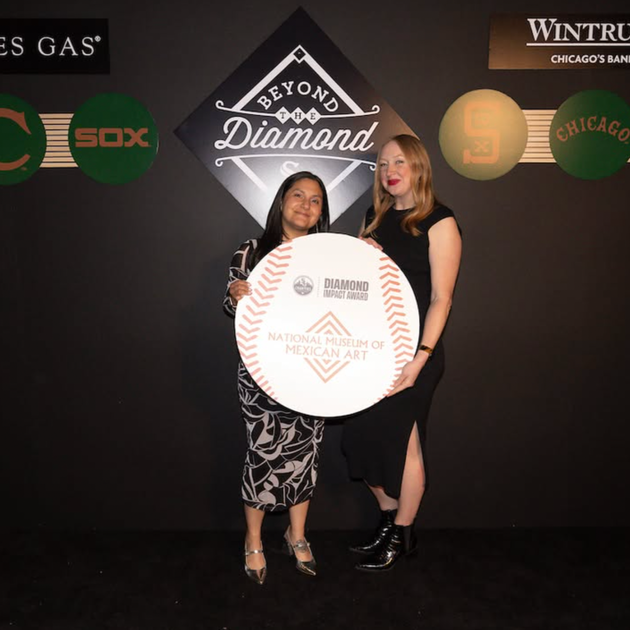 Two women standing indoors, one holding a large round sign that reads 'Diamond Impact Award, National Museum of Mexican Art'. Behind them, a black wall features logos including 'Beyond the Diamond', 'Chicago White Sox', and 'Wintrust Chicago's Bank'.