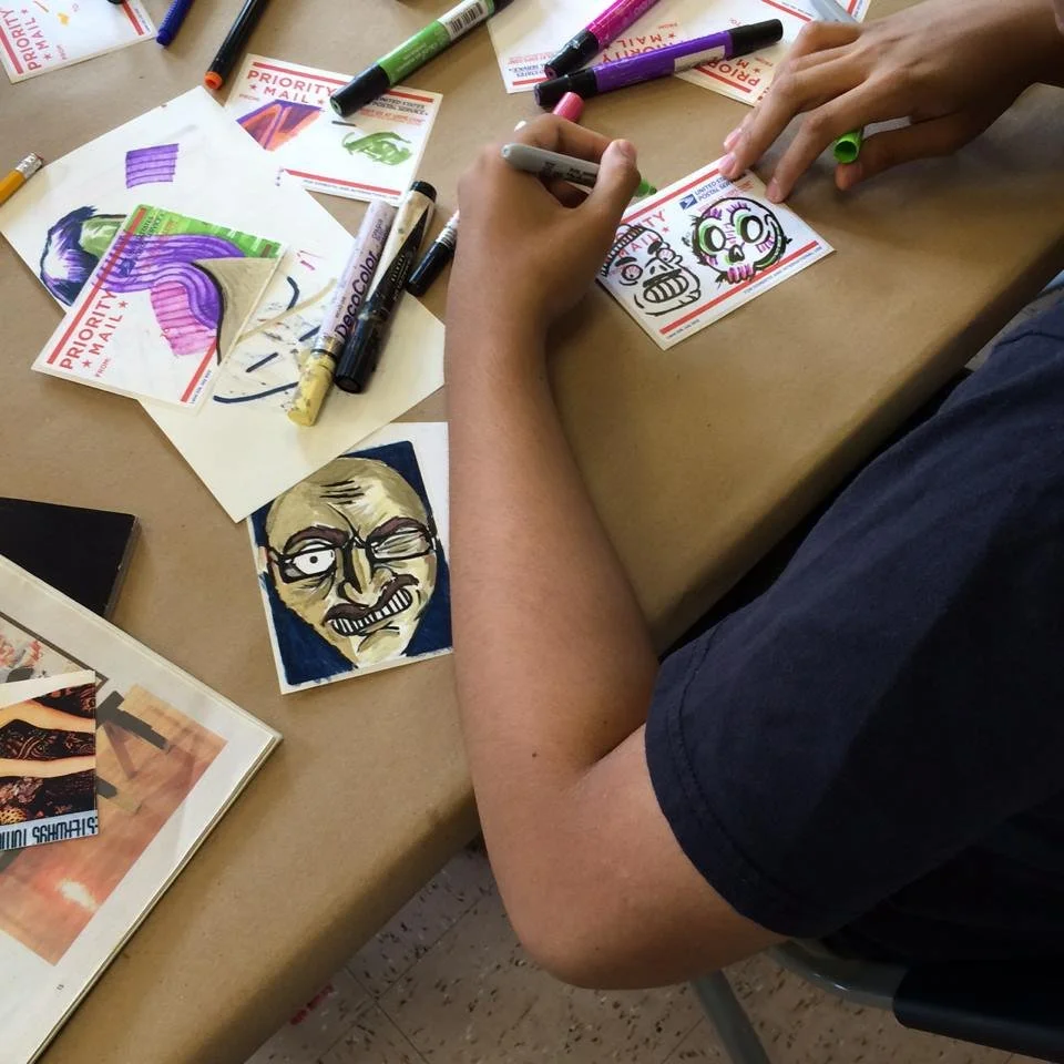 Person drawing colorful, cartoon-style faces on small paper cards with markers, surrounded by various markers and drawings.