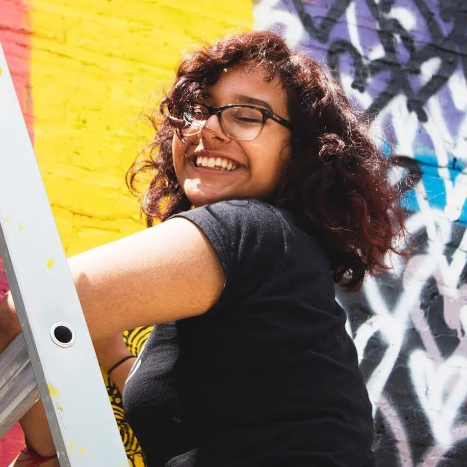 A young person with glasses smiles as they stand on a ladder, behind them is a colorful mural.