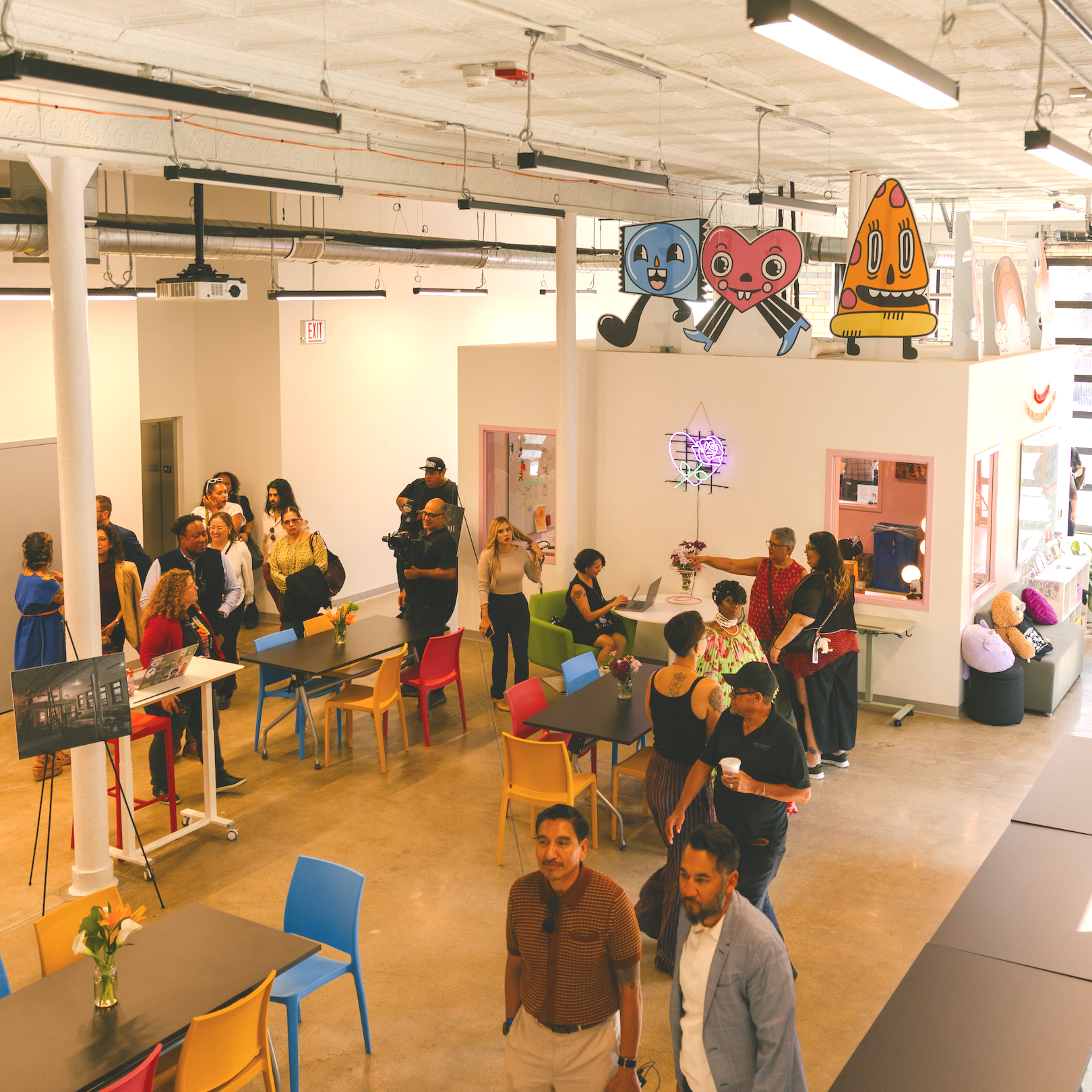 Interior of the Yollocalli firehouse. Multiple groups of people gather at tables or stand, in conversations. Several colorful character standees, chairs, and pillows bring color to the space.