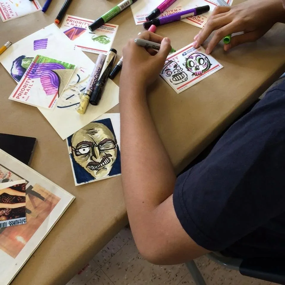 Person creating hand-drawn, cartoon-style sketched faces on USPS priority mail postcards with markers on a table filled with art supplies, including colored markers, printed pictures, and sketchbooks.