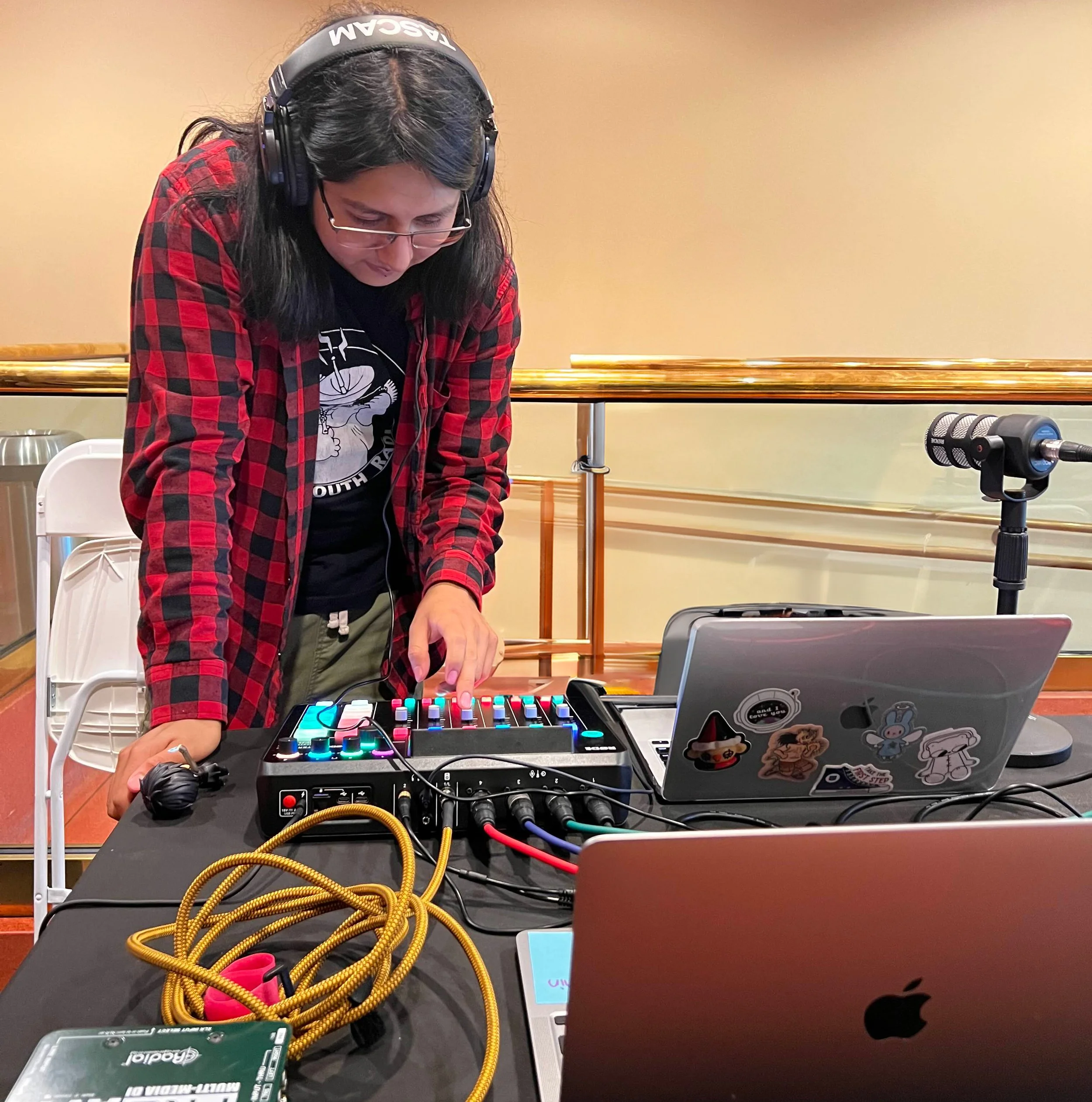 A person with shoulder-length hair and glasses leans over audio equipment. They are wearing a red and black flannel shirt, and a black graphic tee.