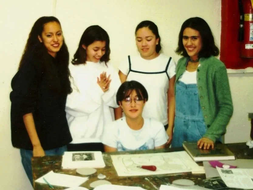 Five young people gather around a table of art supplied. Some are smiling, and others are focused on the work in progress on the table.