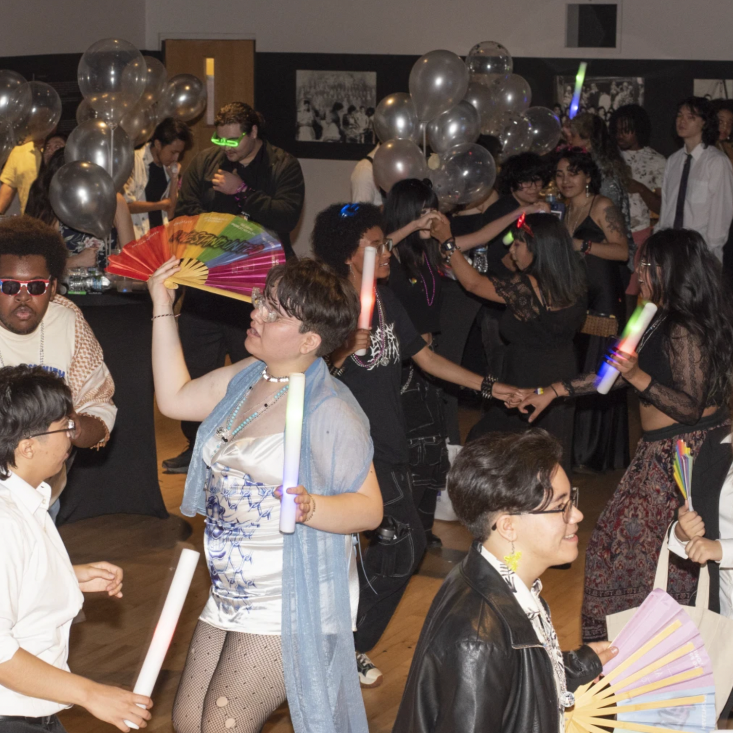 Young people dance on a dimly lit dancefloor. Some people dance in pairs, and some carry fans and glow sticks. Balloons and tables of snacks are visible in the background.