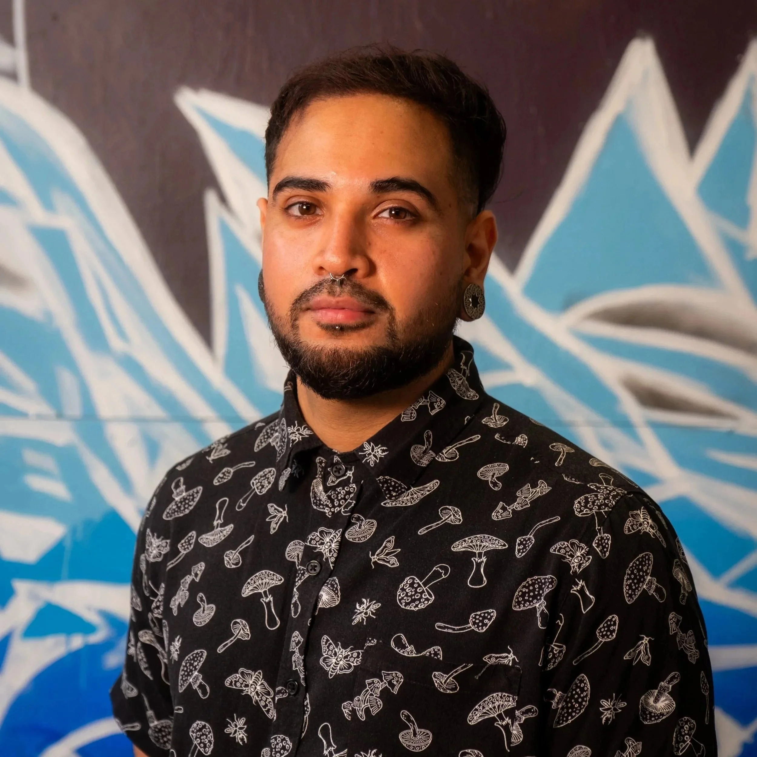 A person with a trimmed beard and gauged ears stands in front of a wall with blue and white graffiti lettering. They arewearing a black button-up shirt that features a mushroom and bug print.