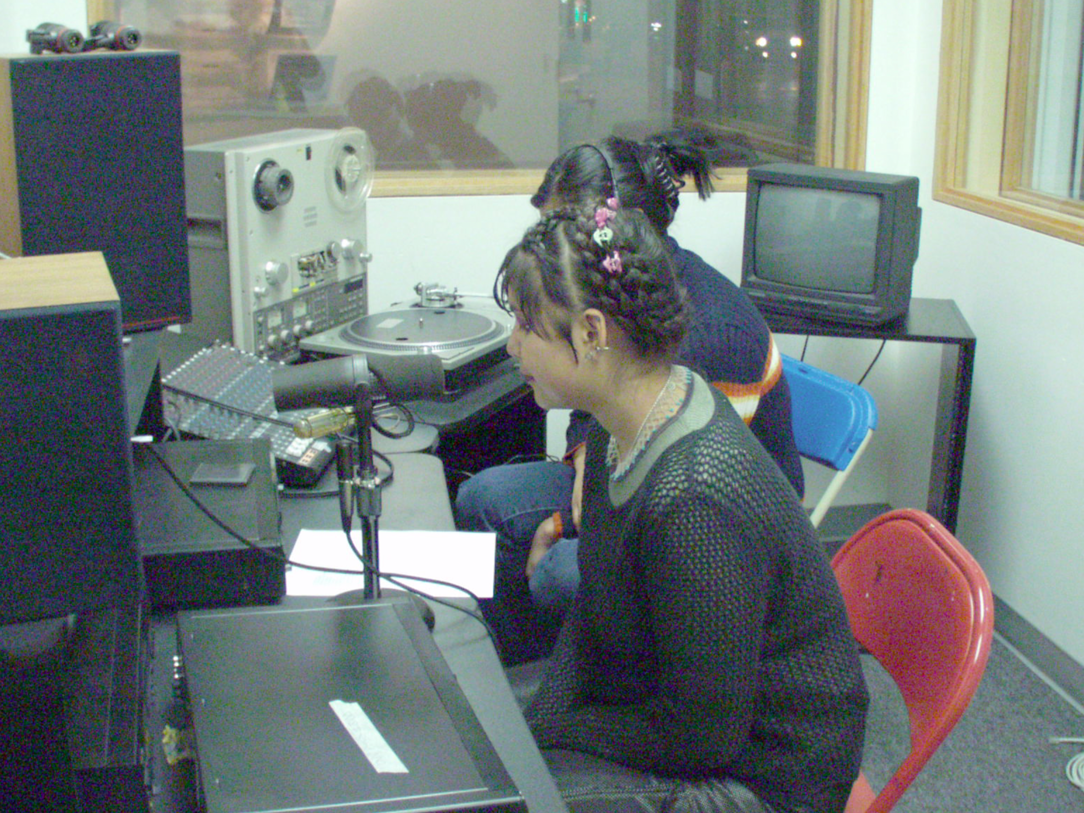 Two young people sit in a radio both and speak into mics. Both have their hair up, in hairclips.