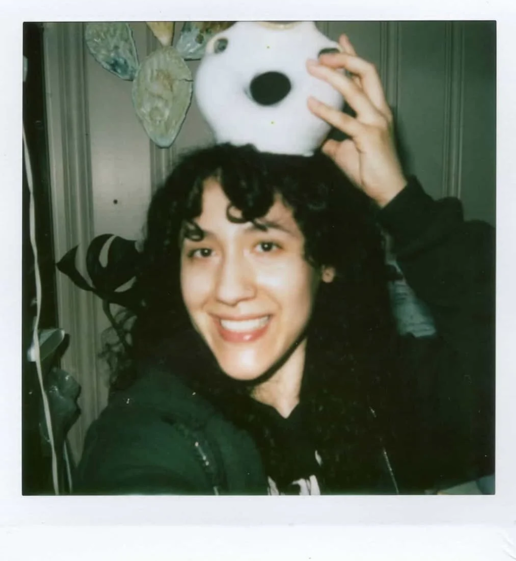 A person with dark curly hair and bangs smiles for a selfie. They are perching a ceramic vase on their head.