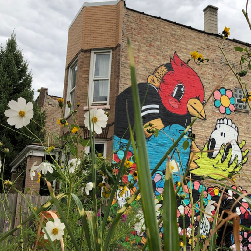 Graffiti mural of a cartoon bird with a red head, and a skeleton with a flower on its head, on the brick wall of a building, with white and yellow flowers in the foreground.