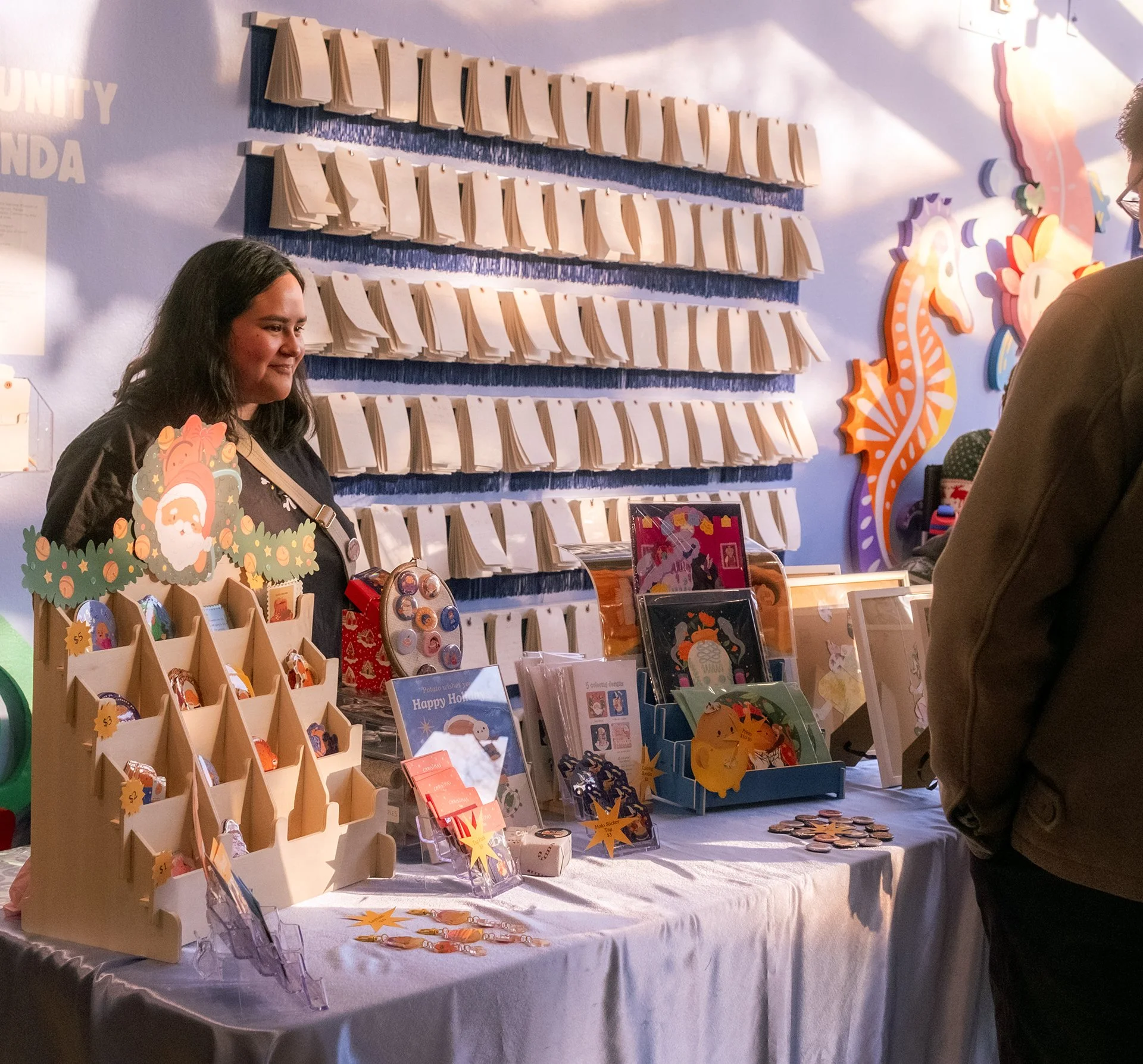 A young person stands behind a table of stickers, art prints, and other goods.