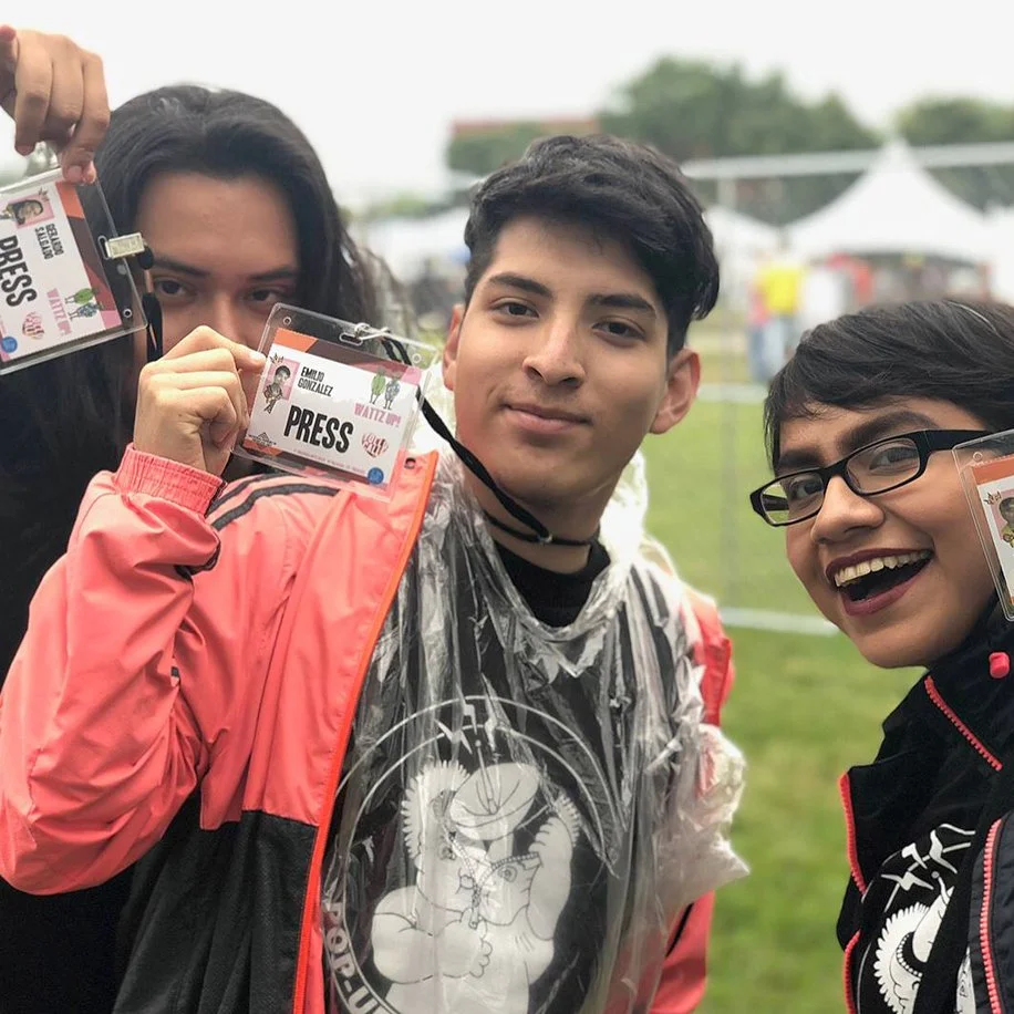 Three young people pose at an outdoor festival. They hold up Press Passes, and wear matching "Pop-up Radio" tee shirts.