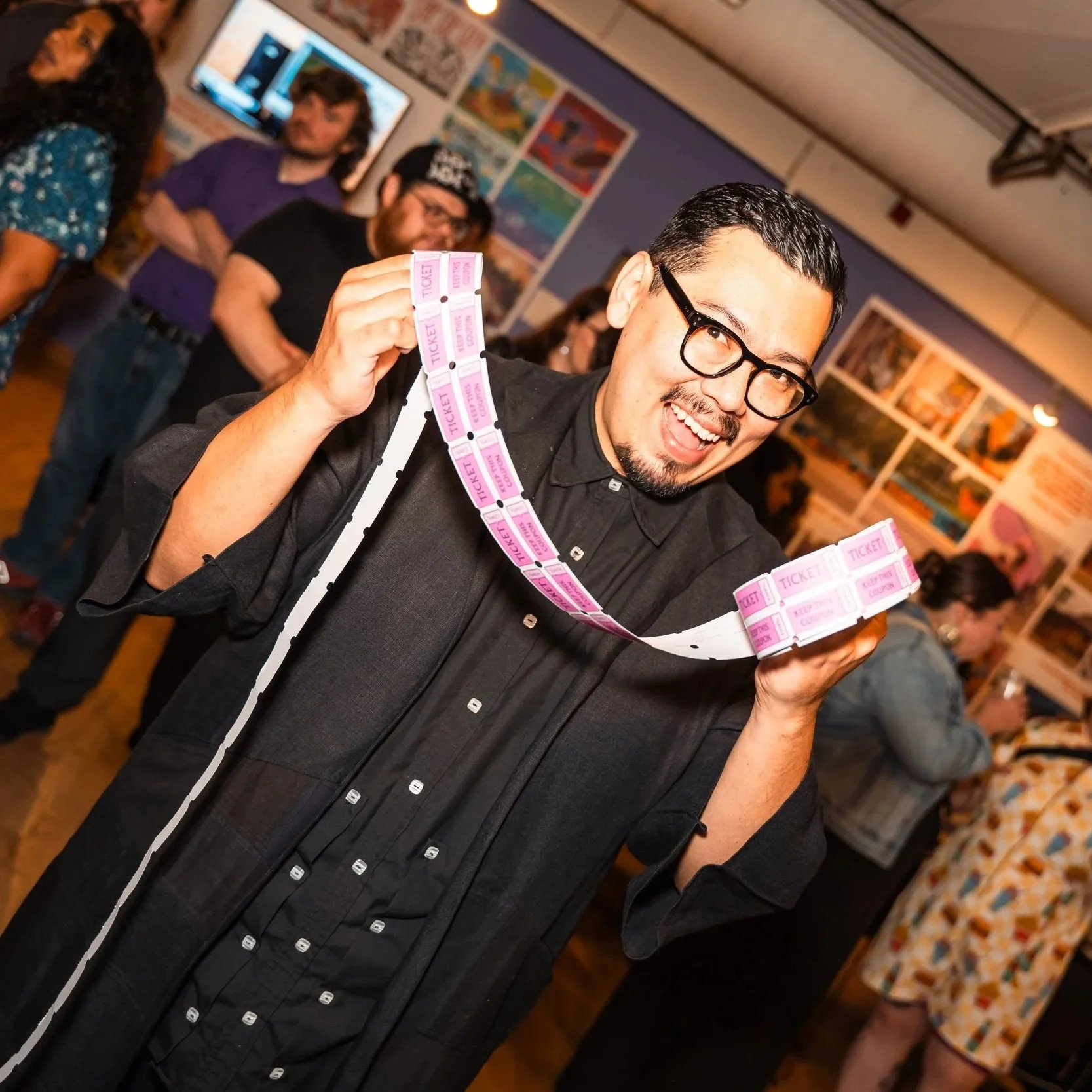 A smiling person in a black guayabera holds up a roll of pink raffle tickets. In the background, people a warmly lit art gallery.
