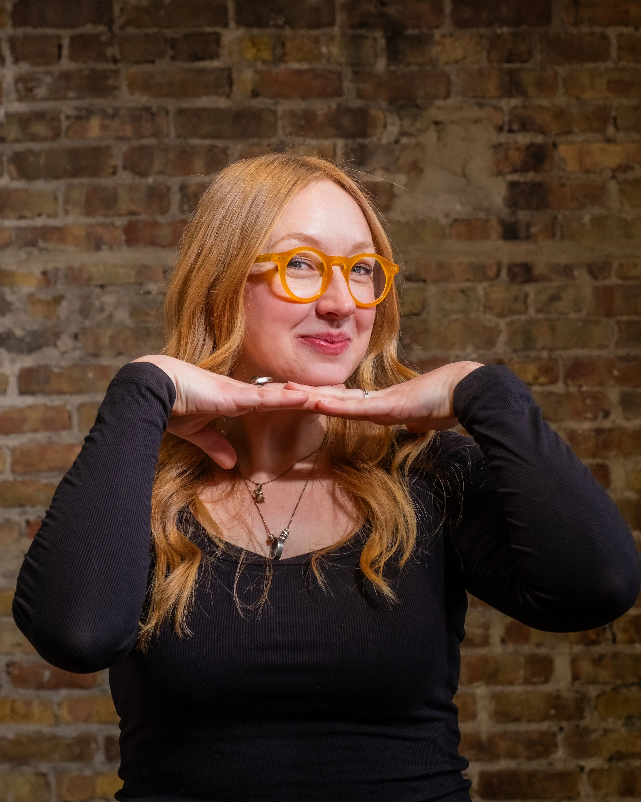 A person with orange glasses and long strawberry blonde hair poses in front of a brick wall. They are smiling and have their head perched on their hands. They are wearing multiple necklaces and rings.