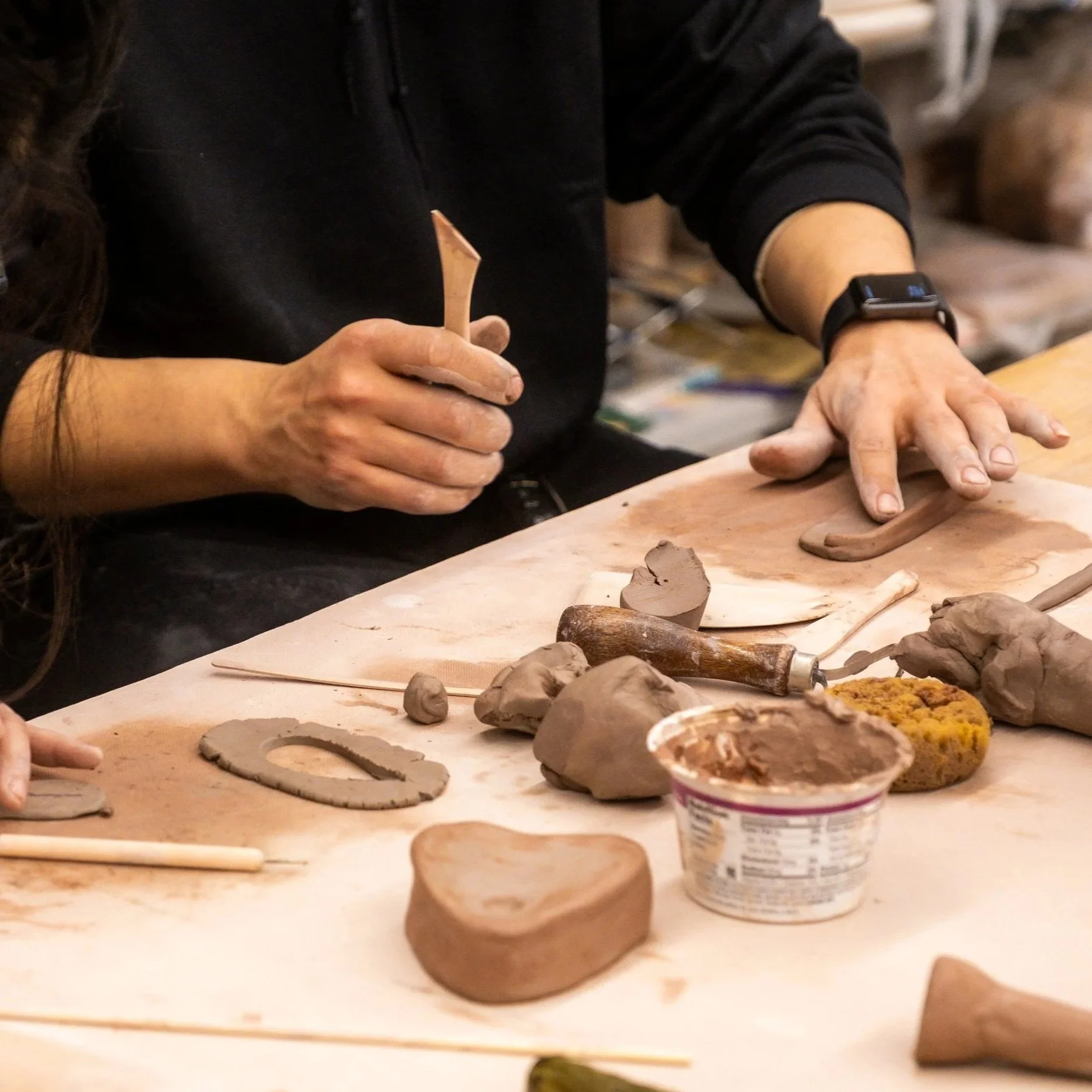Two hands work to shape pieces of brown clay on a wooden table. Various ceramic tools lay on the table.