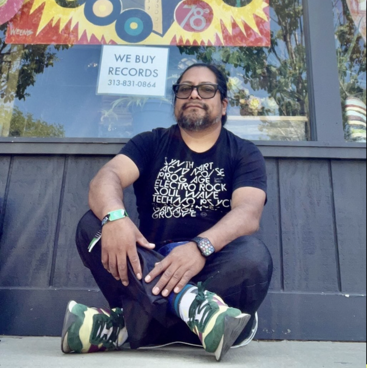 A person with a beard and tinted glasses sits cross-legged in front of a record store. They are wearing a black graffic tee and green and purple sneakers.