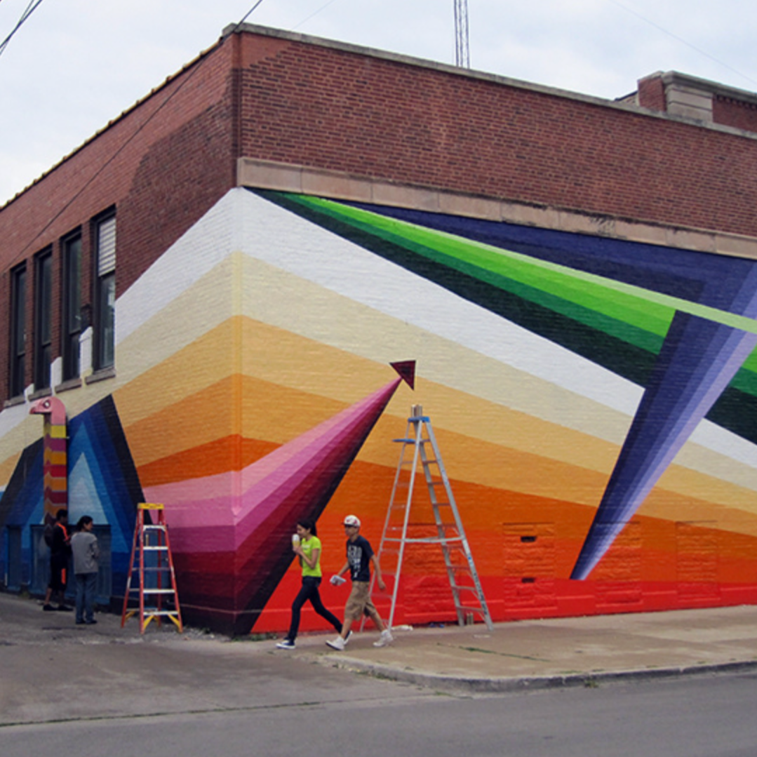 A mural depicting colorful geometric shapes is examened by several young people. Two ladders are set up in front of the mural.
