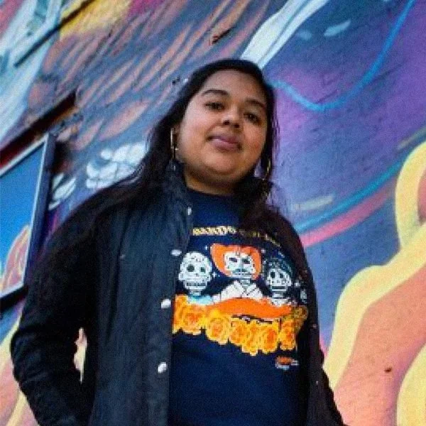 A person with long strait hair poses in front of a mural. They are wearing a dark jacket and gold earings.