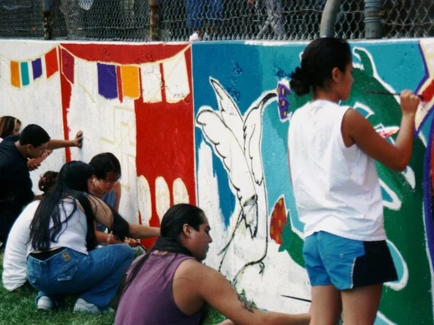 Multiple young people stand and crouch in front of a mural in progress, many hold paint brushes in their hands.