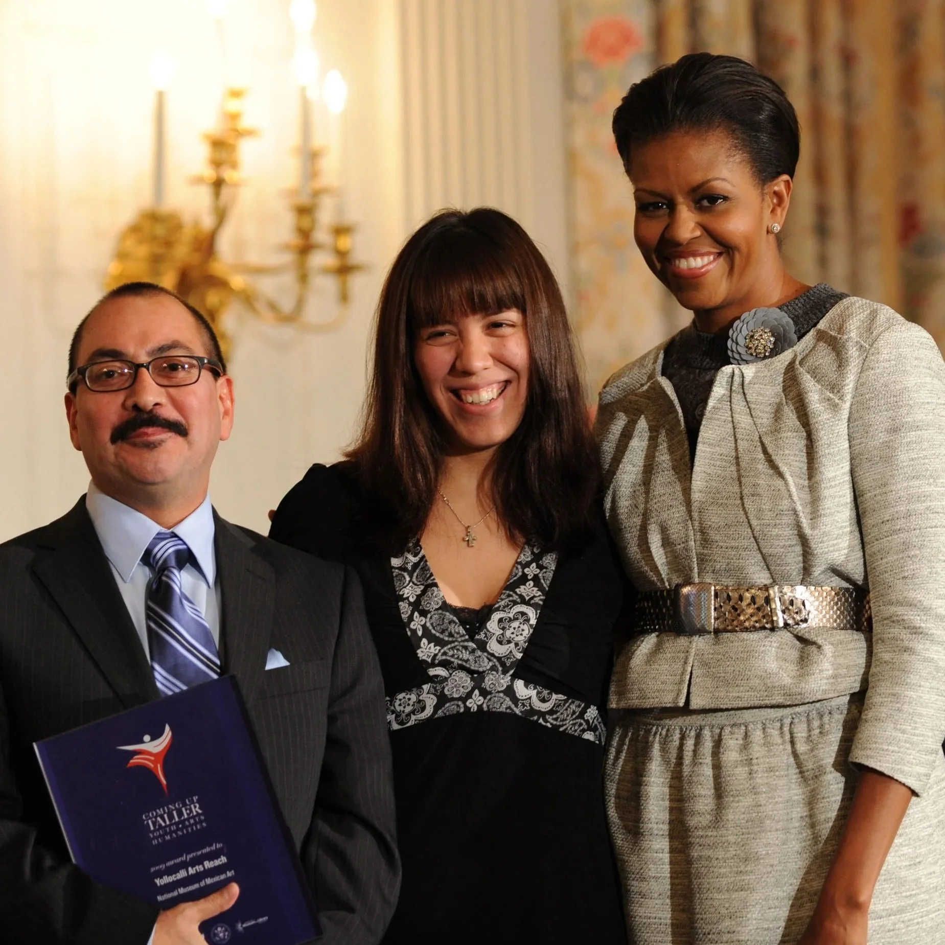 Three people smiling in a formal setting, with a man holding a program, a woman with bangs in the center, and a taller woman with short hair on the right, in front of blurred ornate background and candelabra.