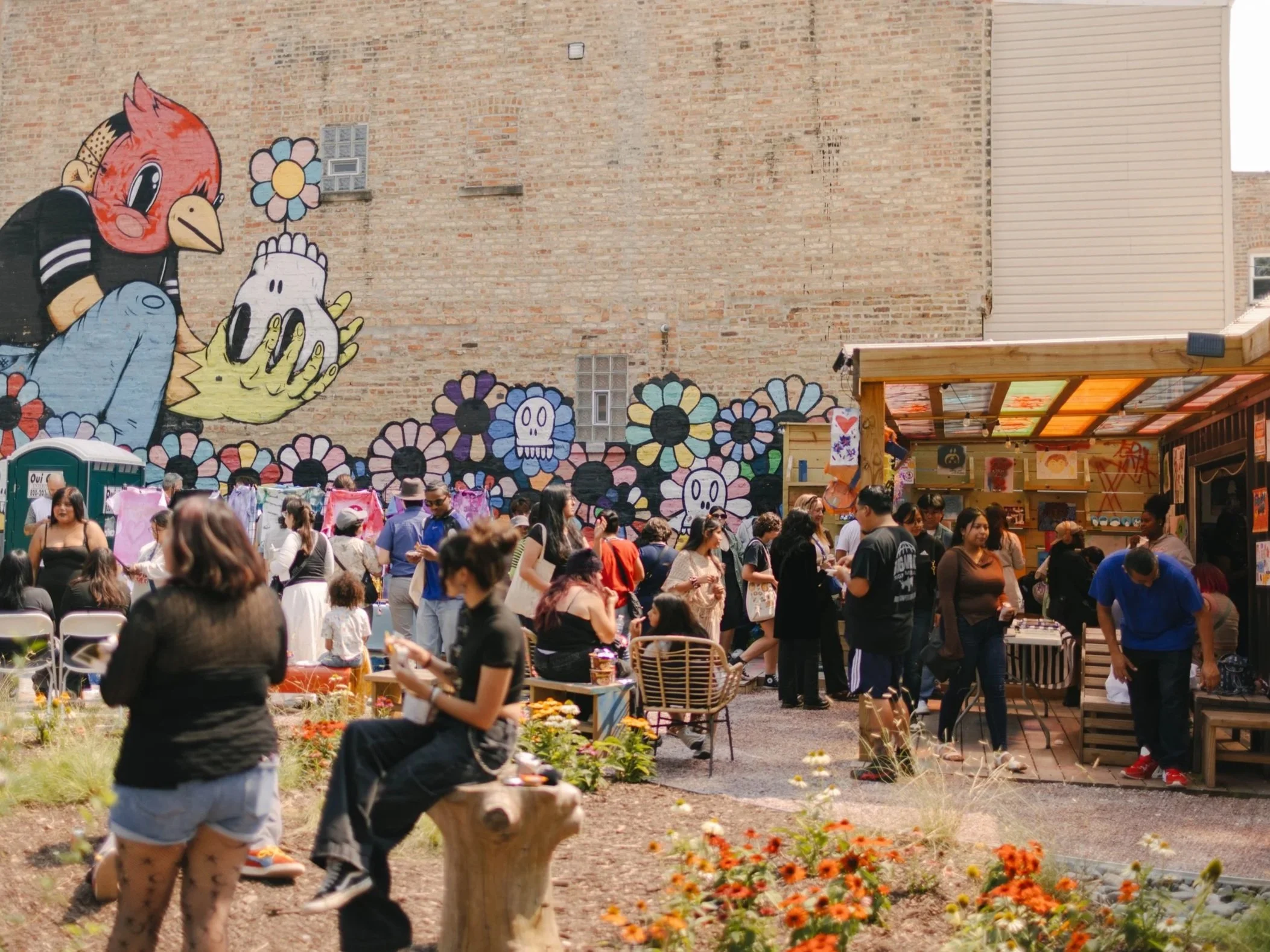 A community gathers at Malinali Garden on a sunny day. Multple people people stand in groups or sit under an awning for shade. A mural depicting a boy in a bird mask holding a skull with a single flower growing out of it can be seen in the background