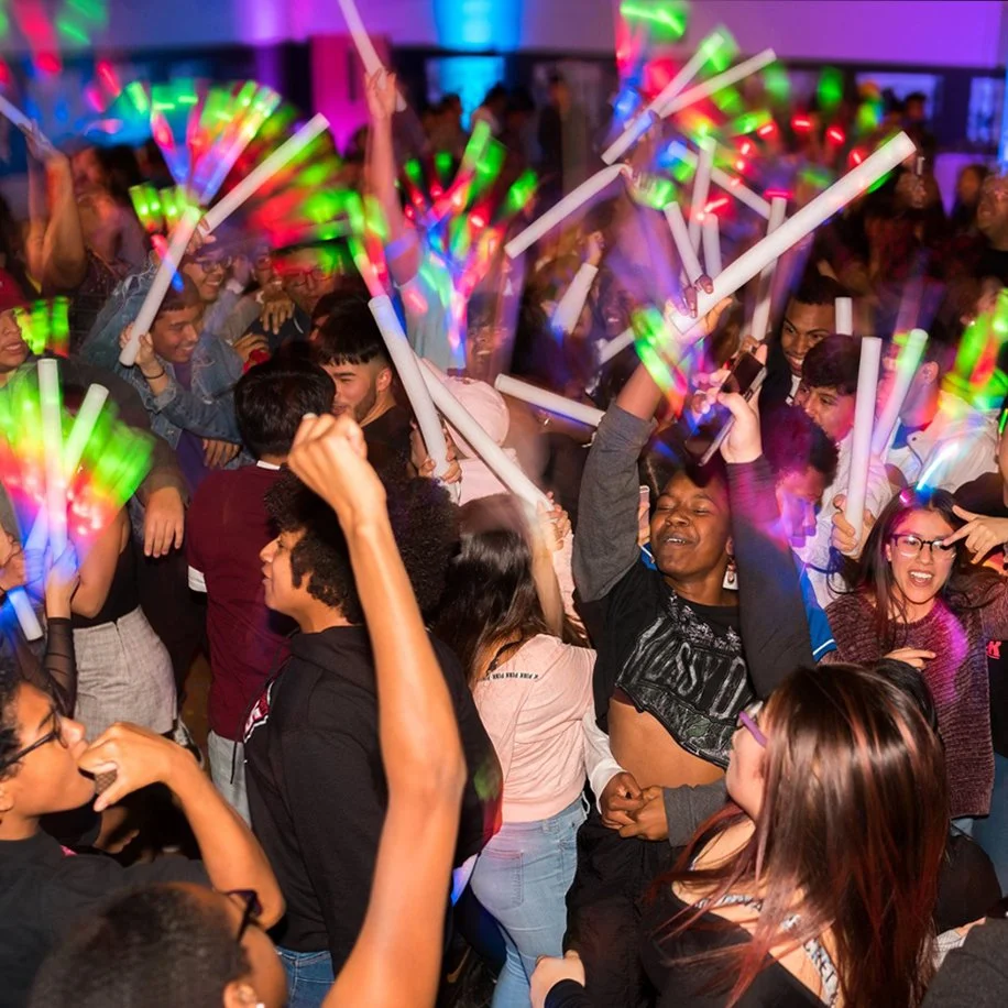 A large group of young people dance on dimly lit dance floor. Glowsticks and other stagelights color the dancefloor.