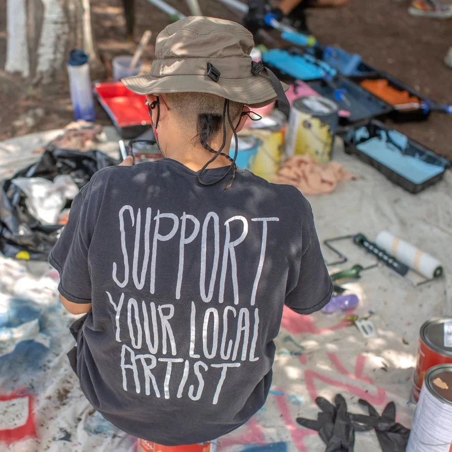 A young artist sits outside, facing away from the camera. They are surrounded by tarps, paint cans and other materials fo an outdoor mural. Their shirt reads "support your local artist."
