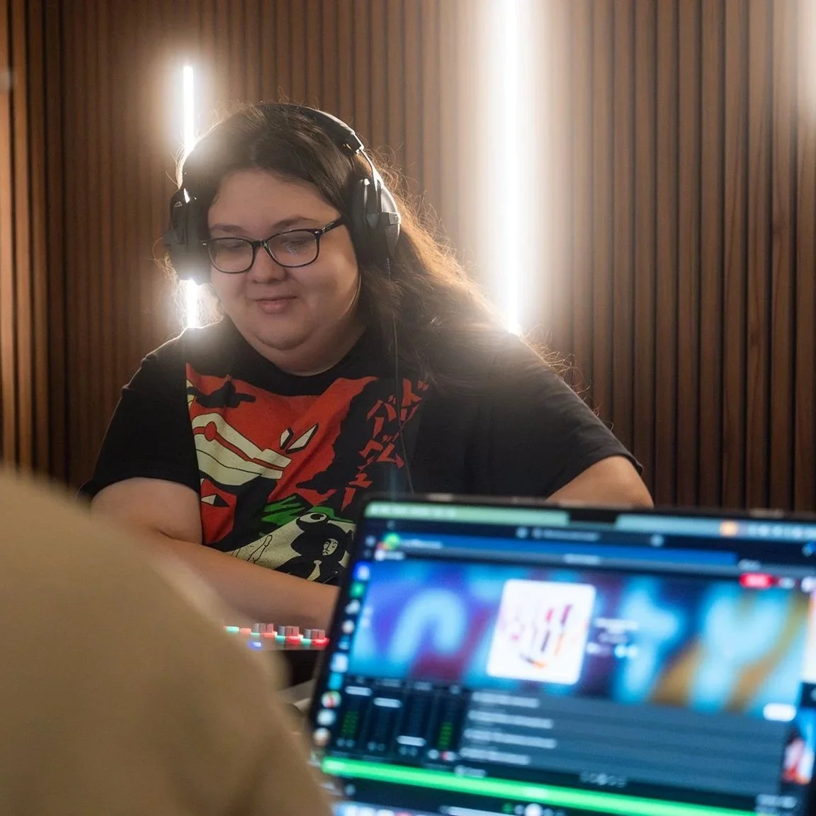 A young person with long hair, glasses, and headphones sits in a fully equiped recording studio. The room is lined with LED lights, and wood panels.