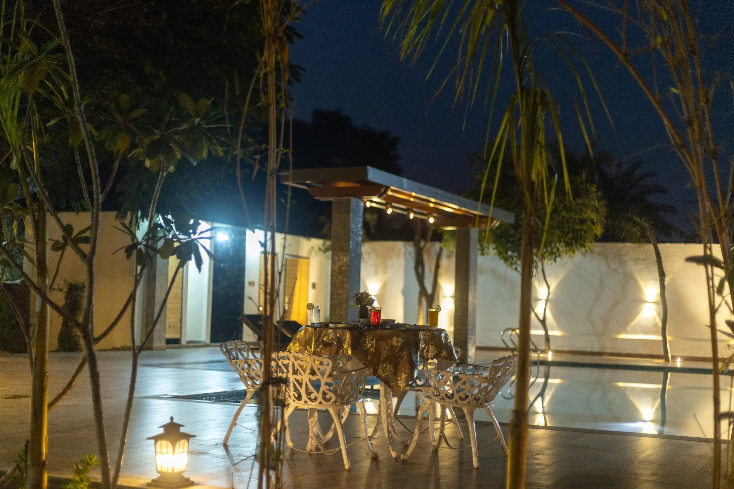Nighttime outdoor patio with a dining table covered with a floral tablecloth, several glasses and a flower vase, surrounded by modern white chairs. There is soft lighting, some trees, and a swimming pool in the background, creating an inviting ambian