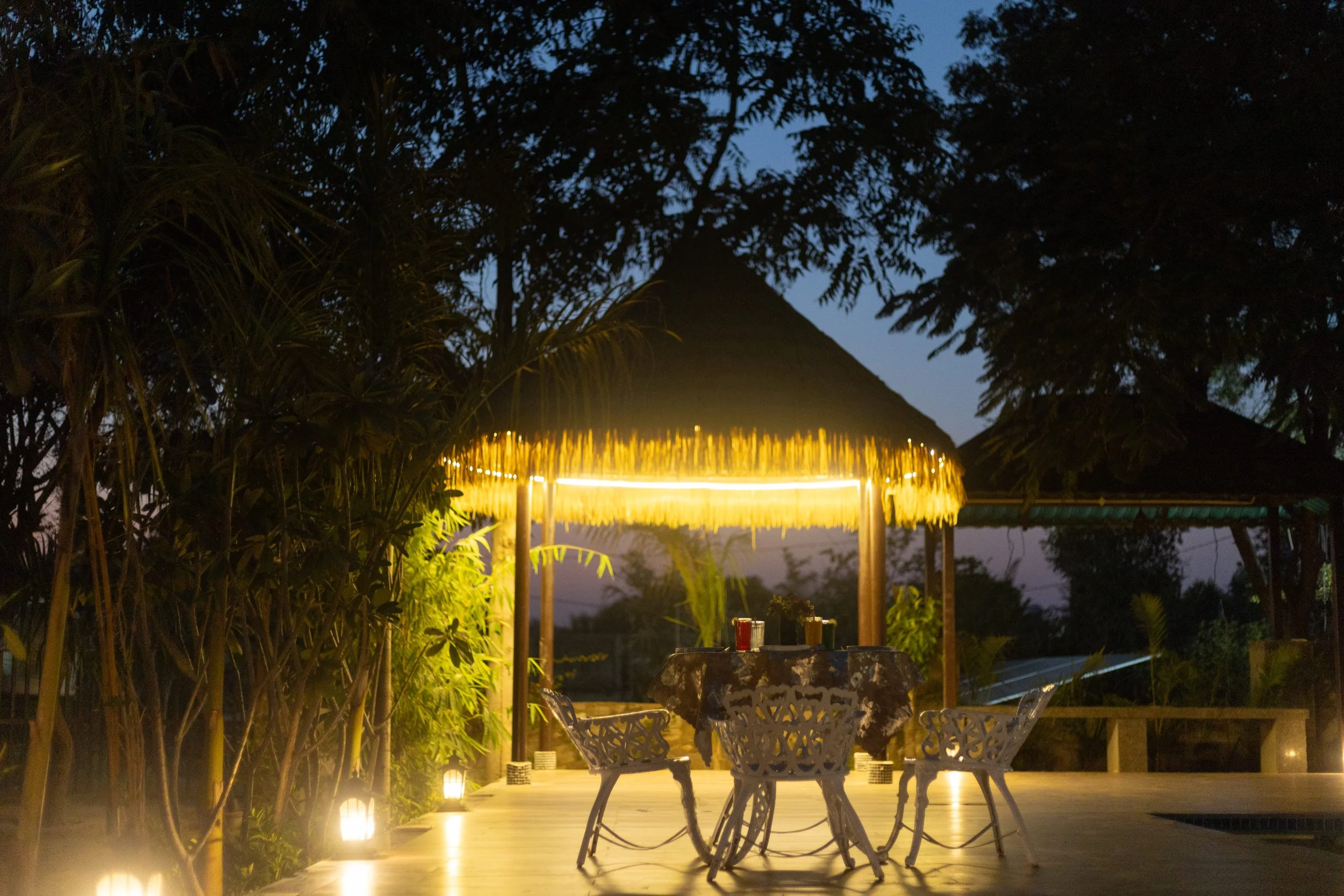 Outdoor dining area at dusk with a thatched-roof hut illuminated by warm lights, surrounded by lush green plants and trees.