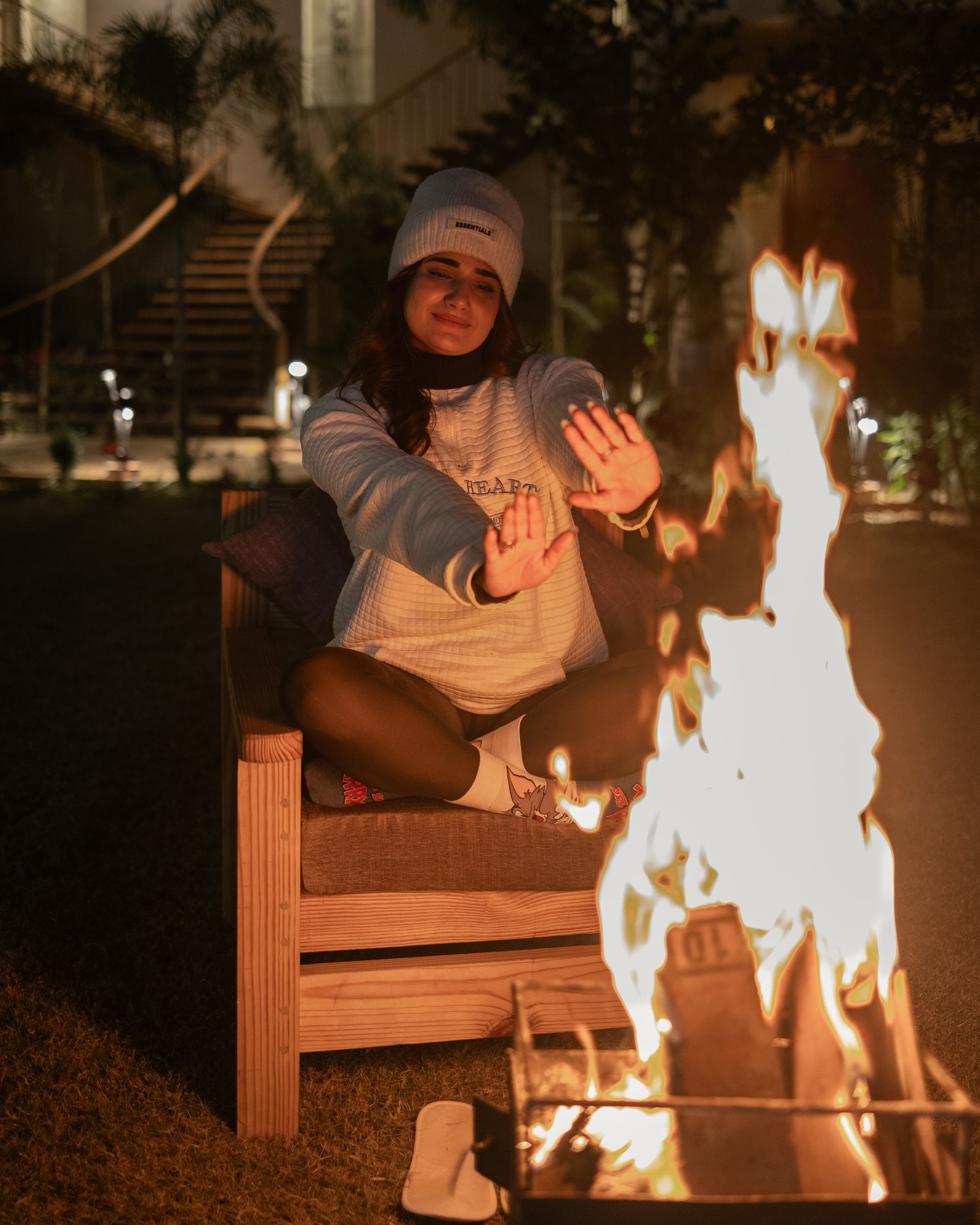 A woman is sitting cross-legged on a bench outdoors at night, wearing a beanie and a long-sleeve shirt, with her hands up as if to shield herself from a large fire in front of her.