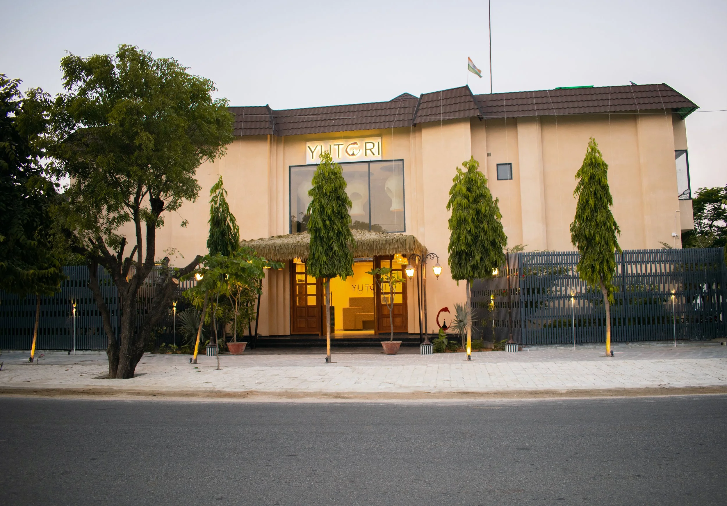A two-story building with a beige facade, multiple green trees in front, a thatched awning over the entrance, and a sign reading 'YUTORI' at the top center. The building is behind a black fence, with a paved sidewalk and road in front.
