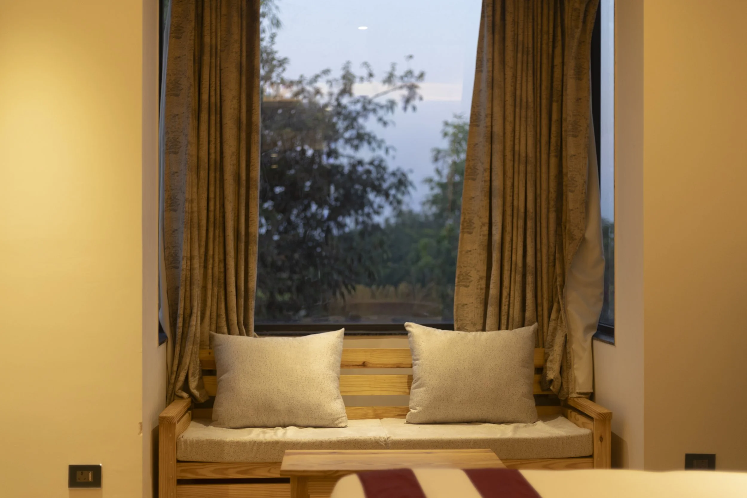 A cozy seating area with a wooden bench and two beige cushions, set against a large window with gold curtains, showing trees and a cloudy sky outside.