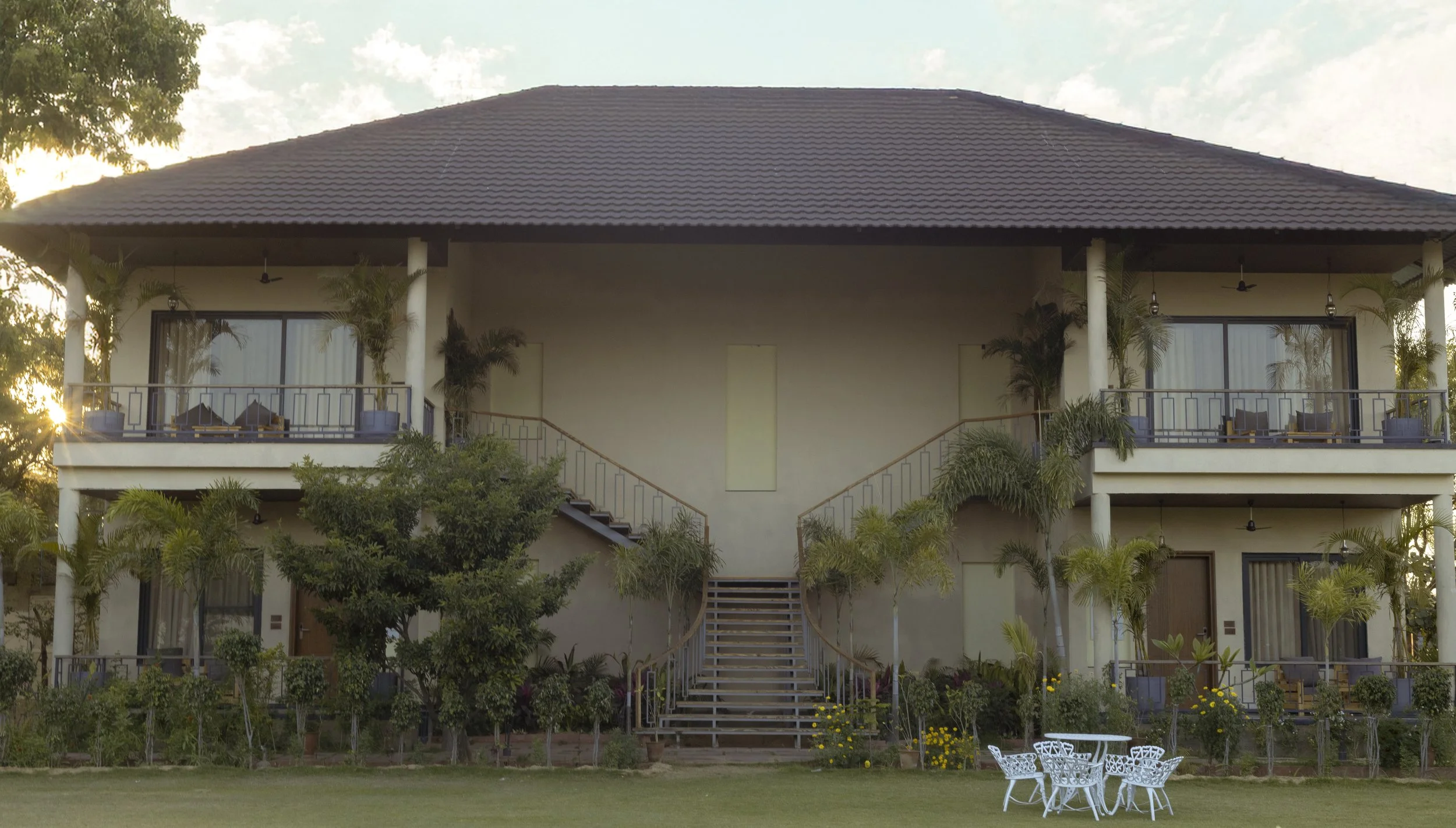 A two-story building with a brown tiled roof, balcony on the right, surrounded by trees and greenery, with a well-maintained lawn in front.