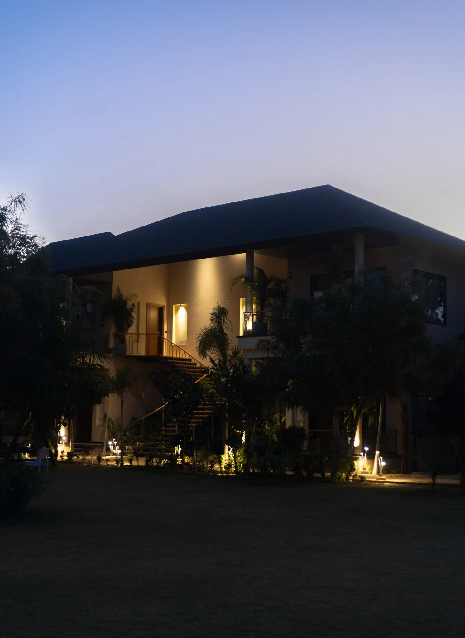 A two-story house illuminated with outdoor lighting, surrounded by trees and bushes, during dusk or early evening.